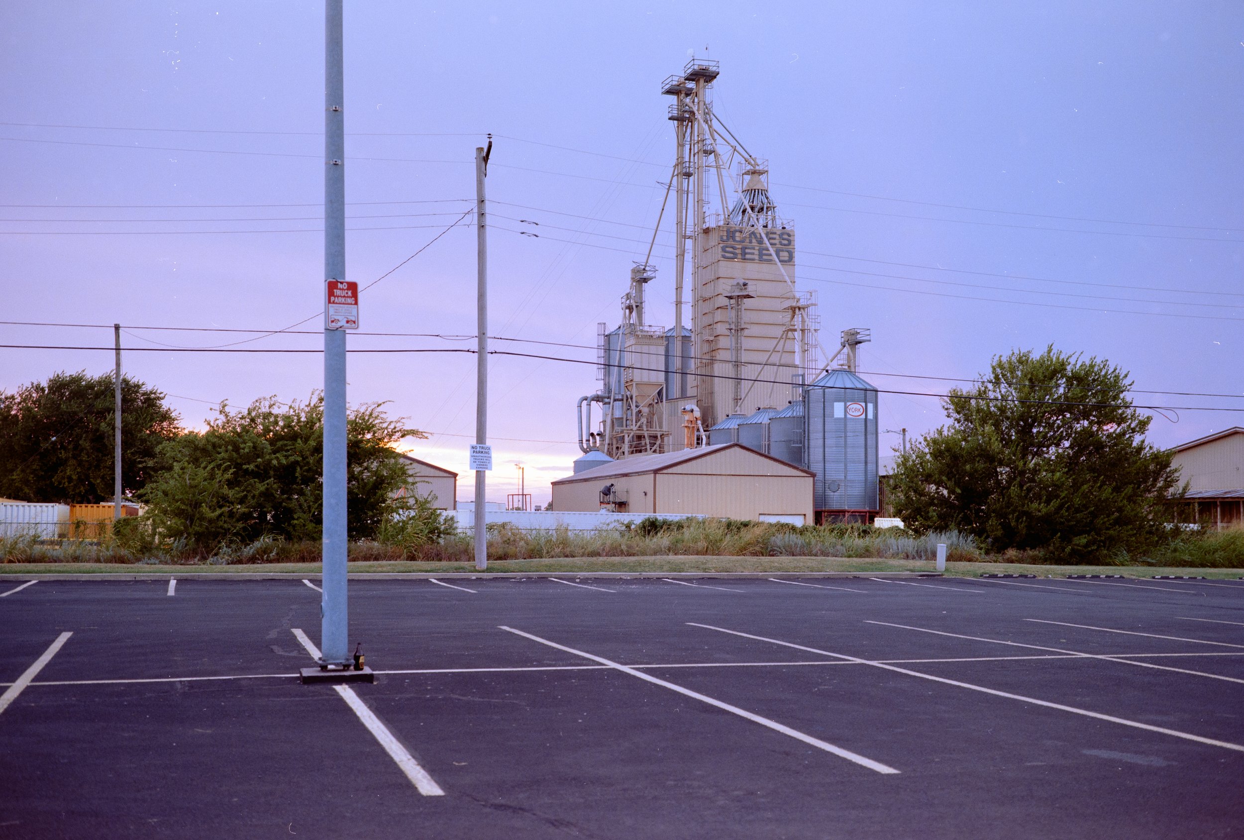 Un parcheggio vuoto con alcune auto parcheggiate ai lati e un vecchio complesso industriale con silos e strutture metalliche sullo sfondo.