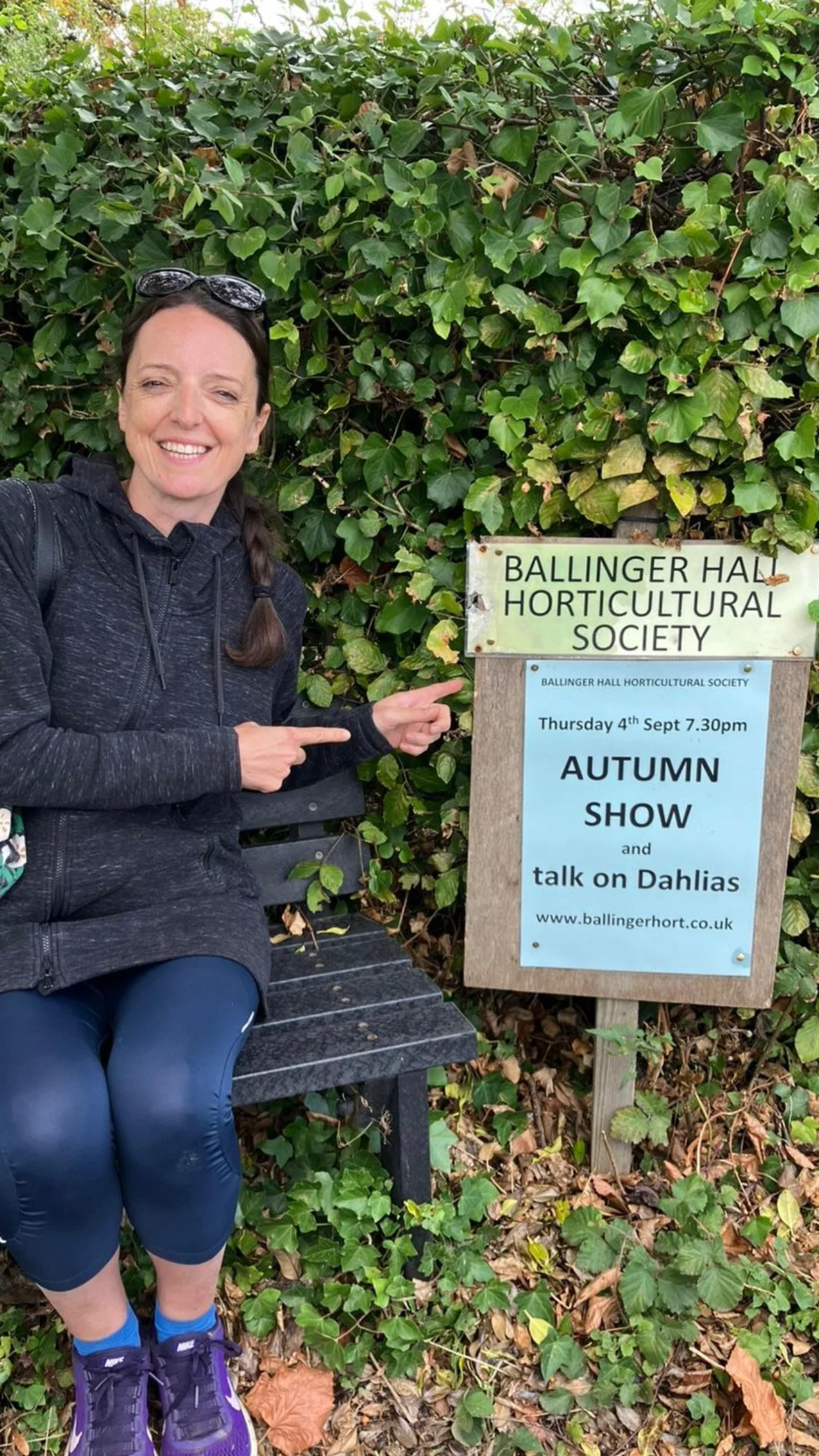 A woman sitting on a bench pointing at a sign for an autumn show and talk on Dahlias organized by the Ballinger Hall Horticultural Society, in front of a leafy green hedge.