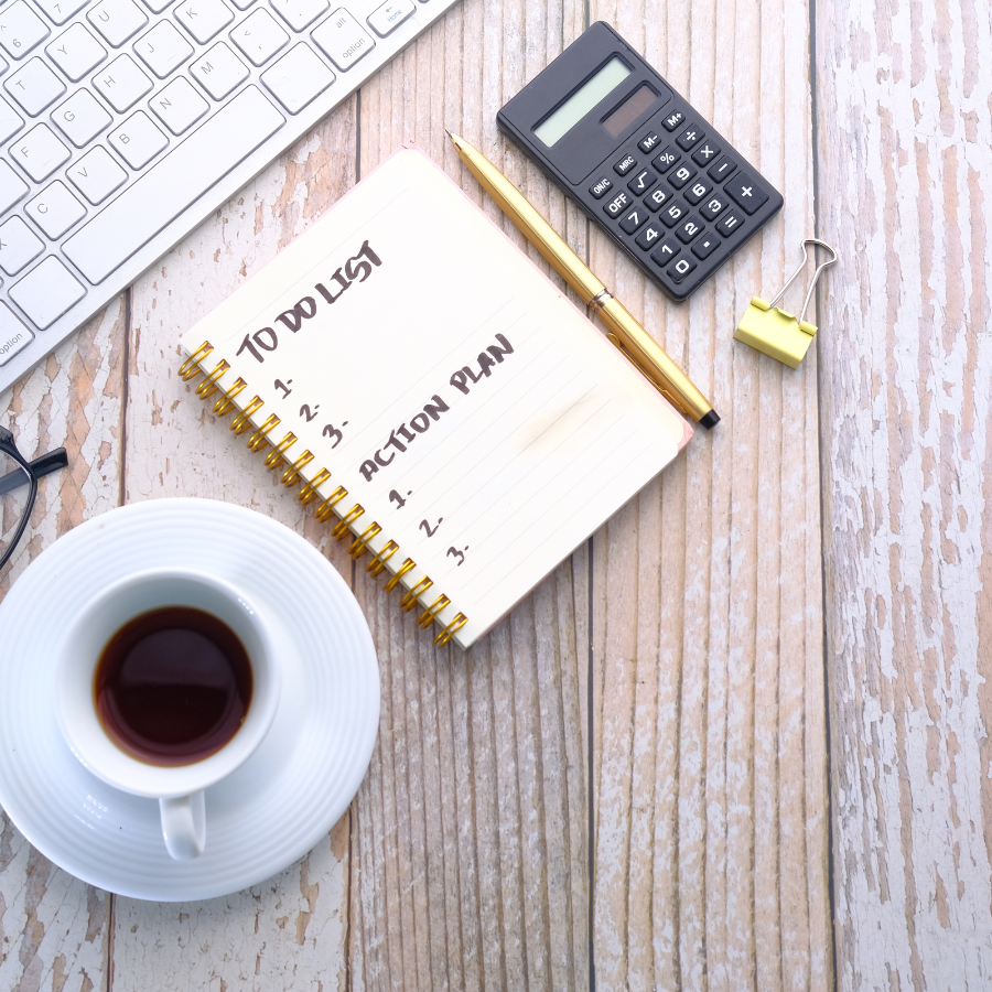 A workspace with a coffee cup, a planner listing to-do and action plan items, a gold pen, a calculator, a binder clip, a pair of glasses, and part of a white keyboard on a wooden surface.