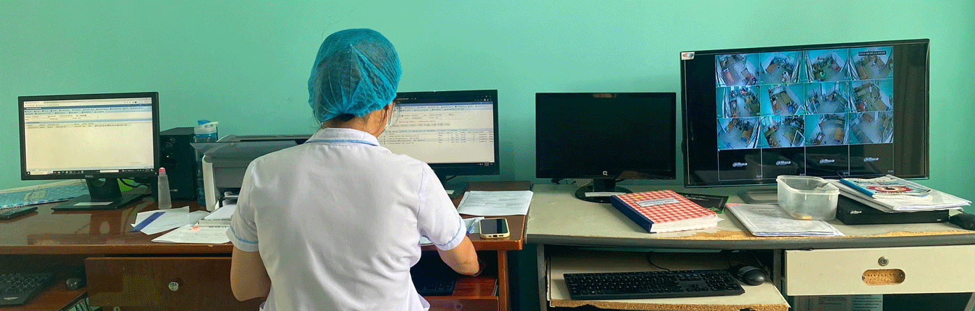 A health worker at a desk reviewing monitoring screens and paperwork in a clinic office