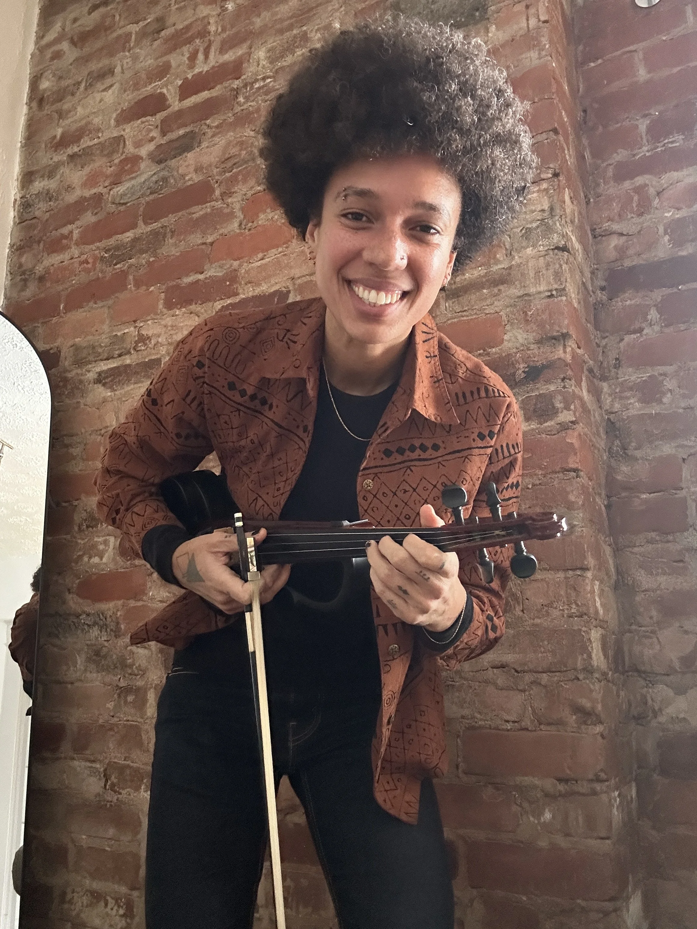 A woman with curly hair smiling and holding a violin, standing in front of a brick wall.