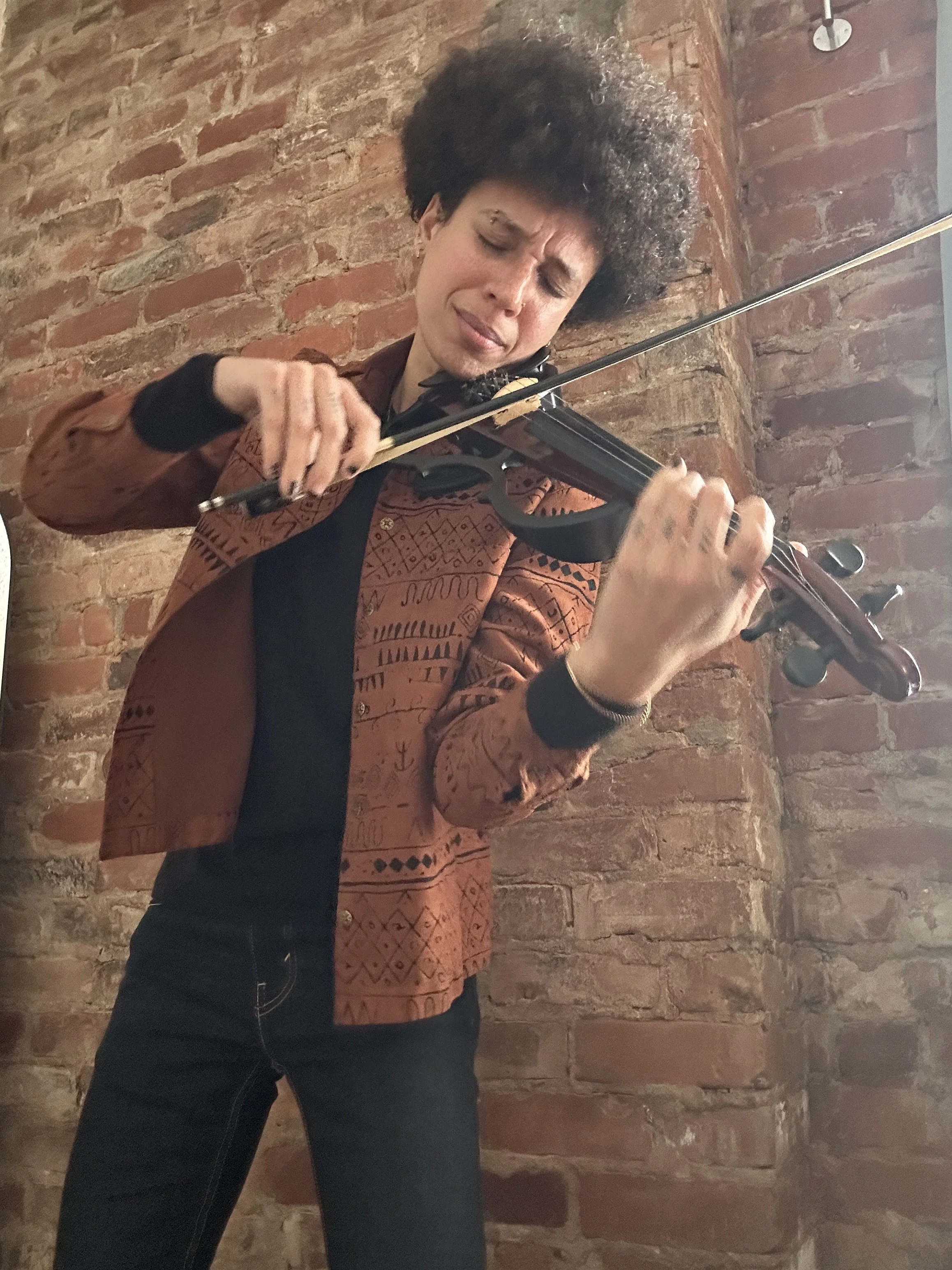 Woman playing a violin against a brick wall, wearing a patterned jacket and black pants.