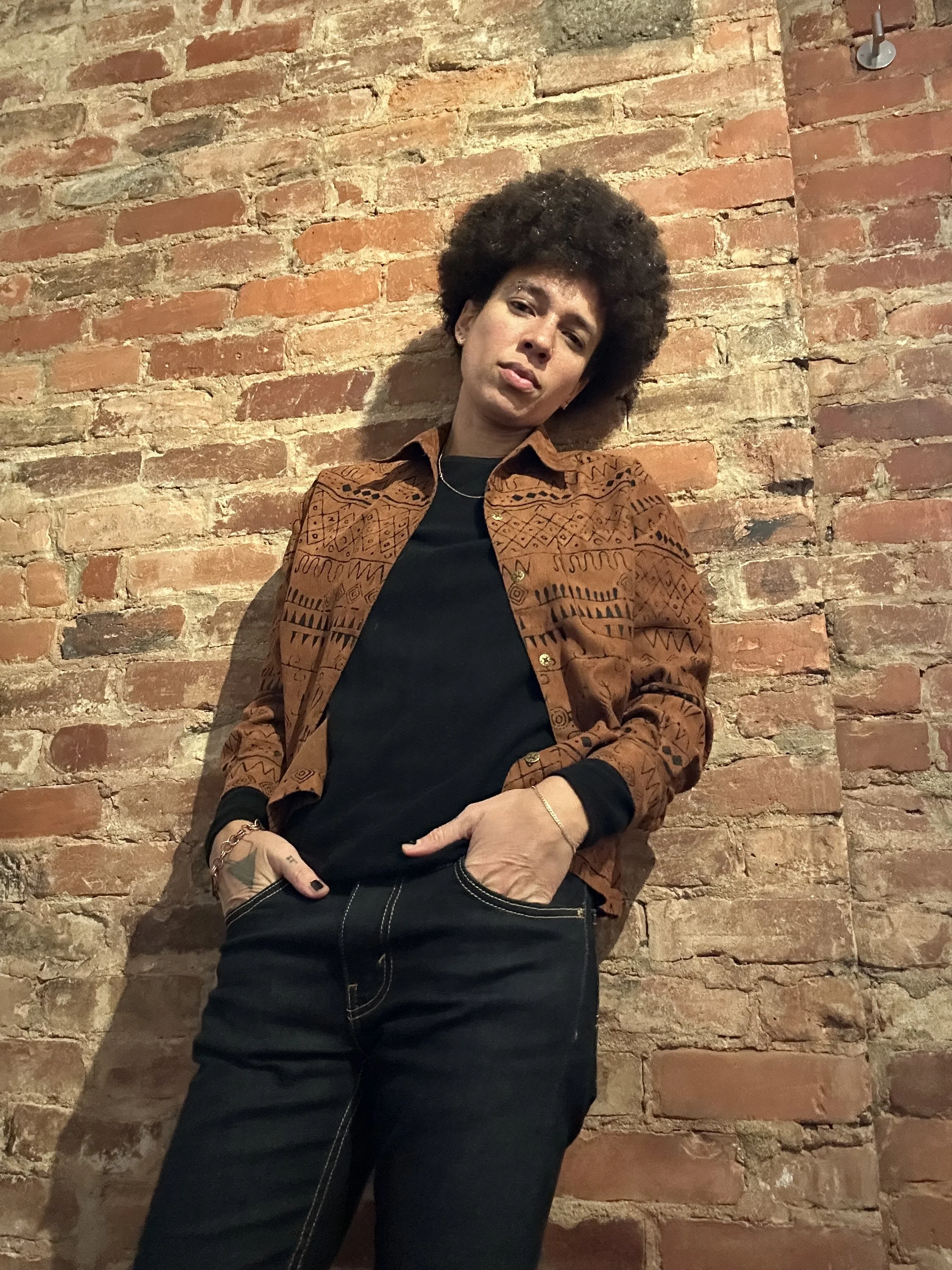 A woman with curly hair standing against a brick wall, wearing a patterned brown jacket over a black shirt and black jeans, with her hands in her pockets and a serious expression.