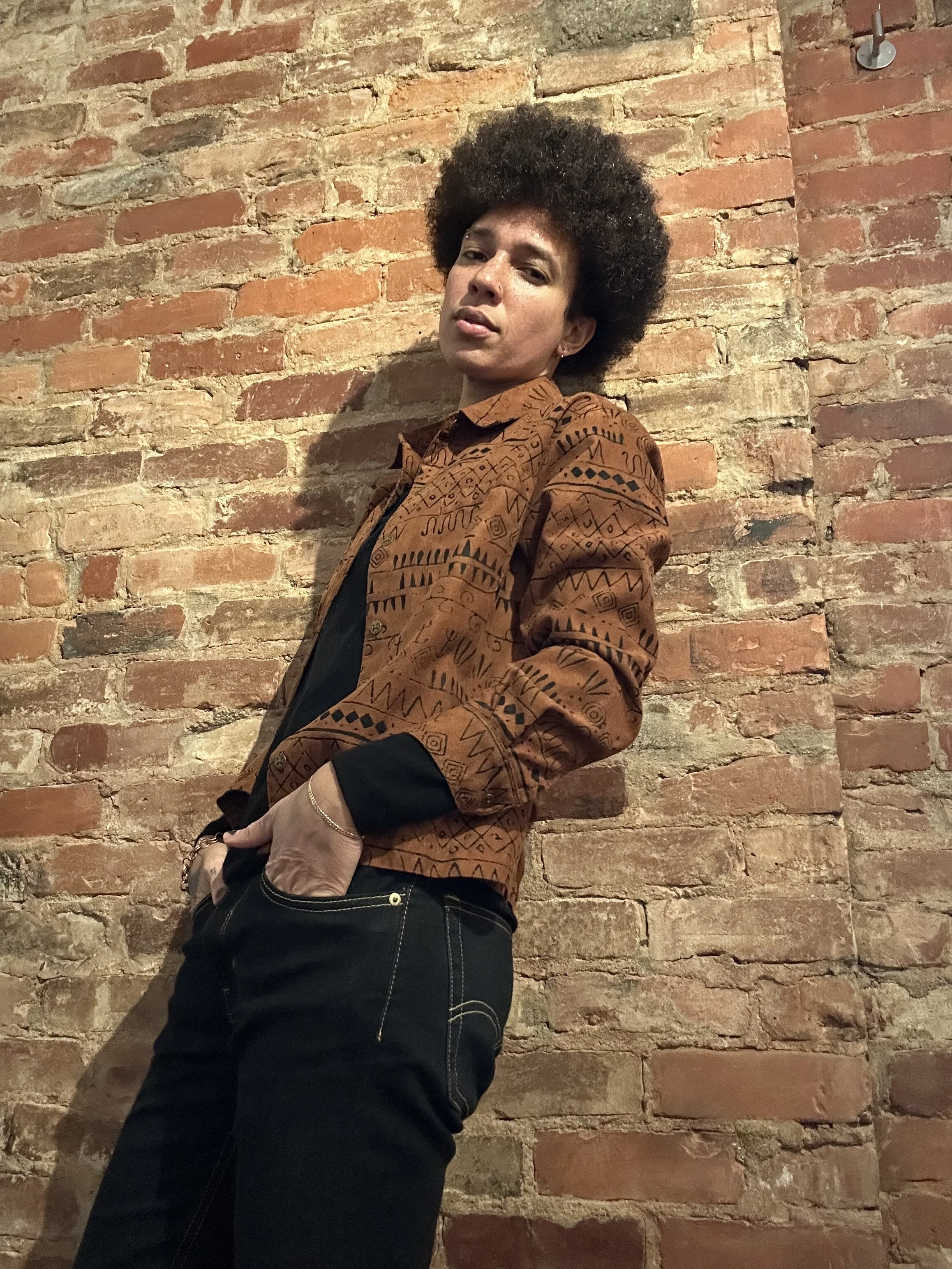 A woman with an afro hairstyle wearing a patterned brown jacket and black jeans standing against a exposed brick wall.