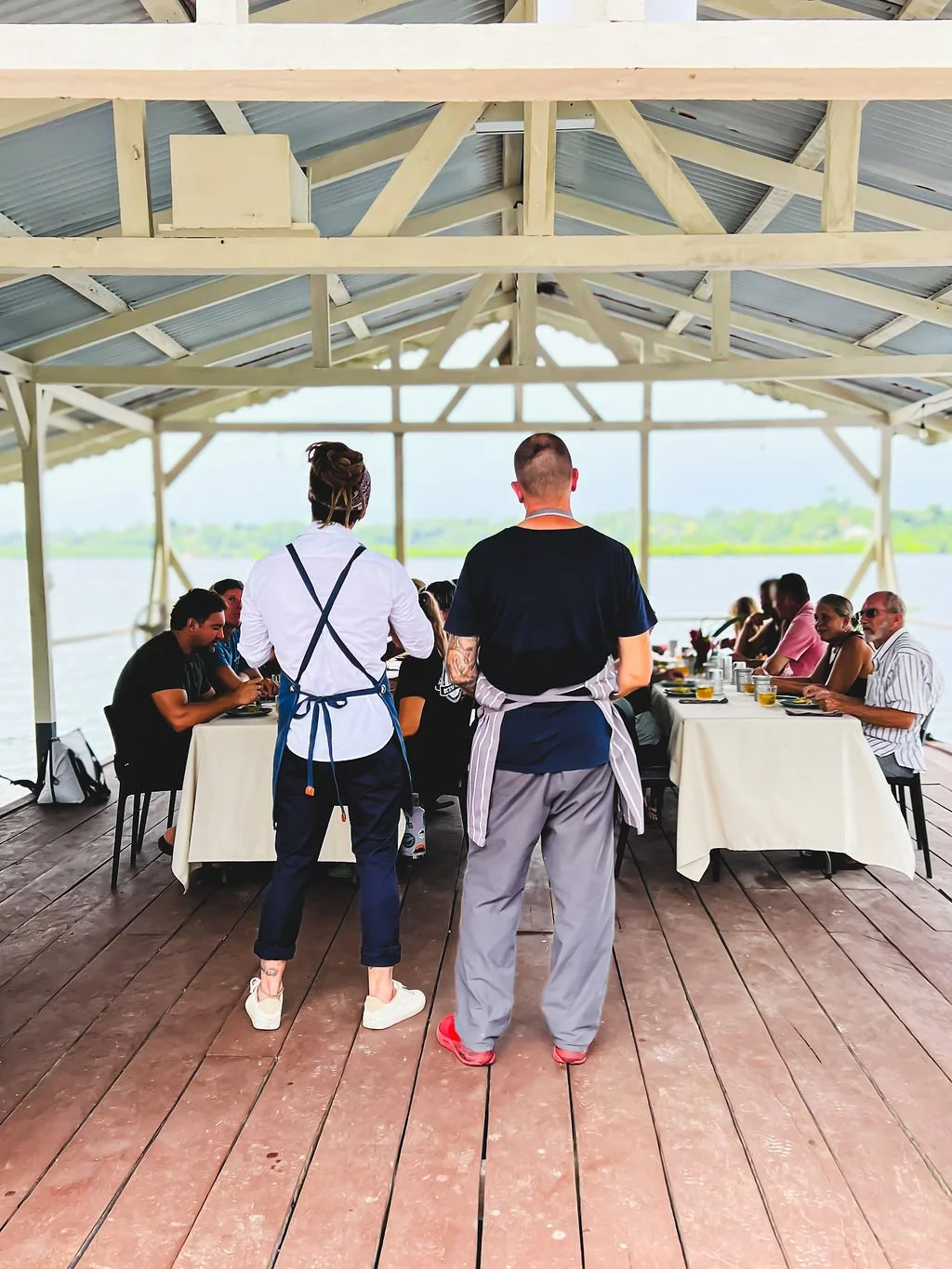 Upscale outdoor dining area by the water with a long table and guests, two servers in aprons stand with their backs to the camera, overlooking guests seated on wooden deck, with a lake and green landscape in the background.