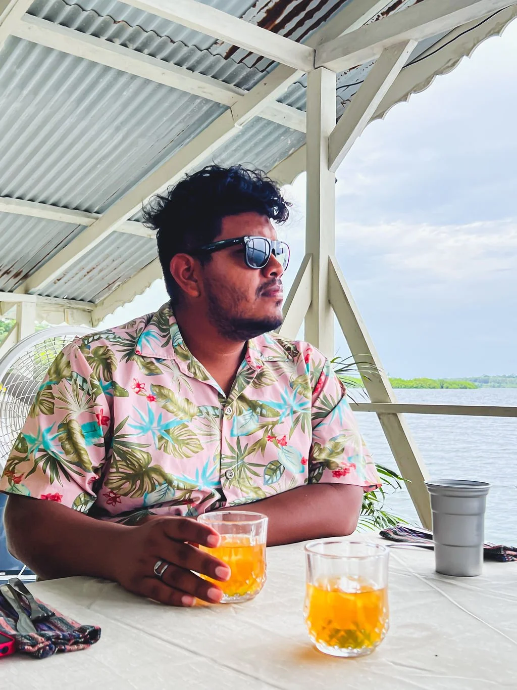 A man wearing a Hawaiian shirt and sunglasses sitting at a table with two glasses of orange drinks outdoors near the water, under a metal-roofed structure.