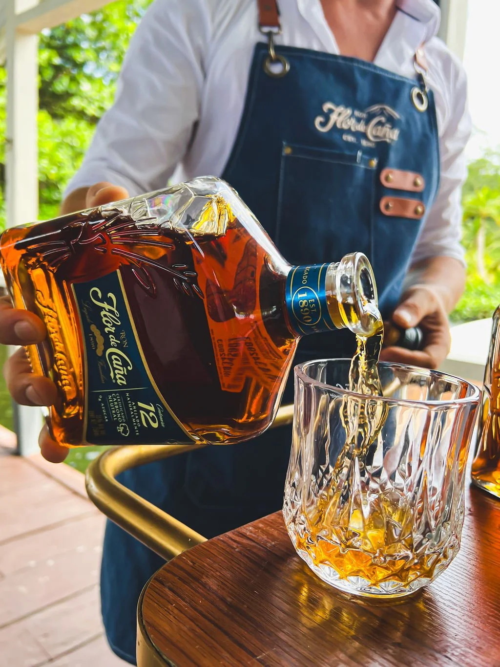 Person pouring rum into a glass on a wooden table, wearing a white shirt and blue apron with 'Farm Girl' embroidered on it, set outdoors with green foliage in the background.