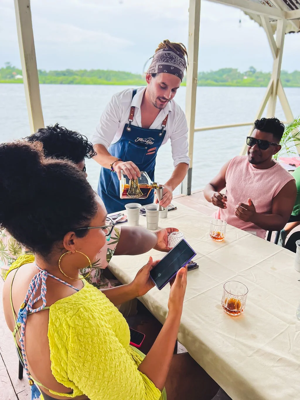 People enjoying drinks at an outdoor waterfront restaurant with a man pouring a beverage into cups and a woman taking a photo, with lush greenery in the background.