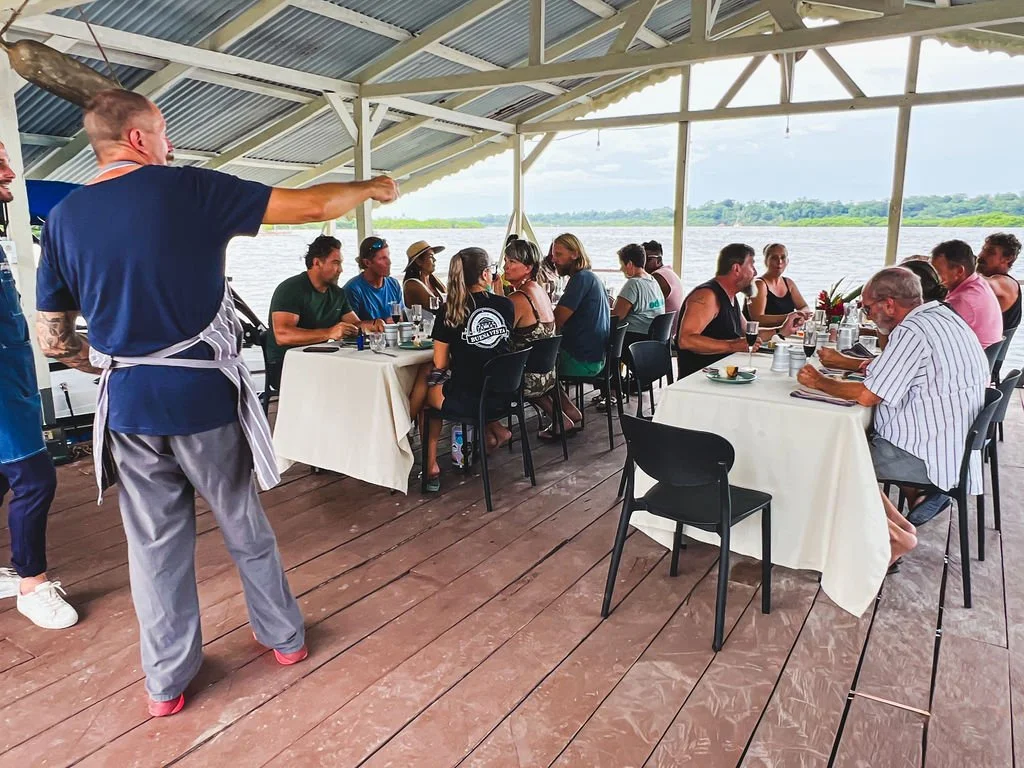 A group of people gathered around tables on a covered outdoor deck overlooking a body of water, with a chef in a blue shirt and apron gesturing while speaking.