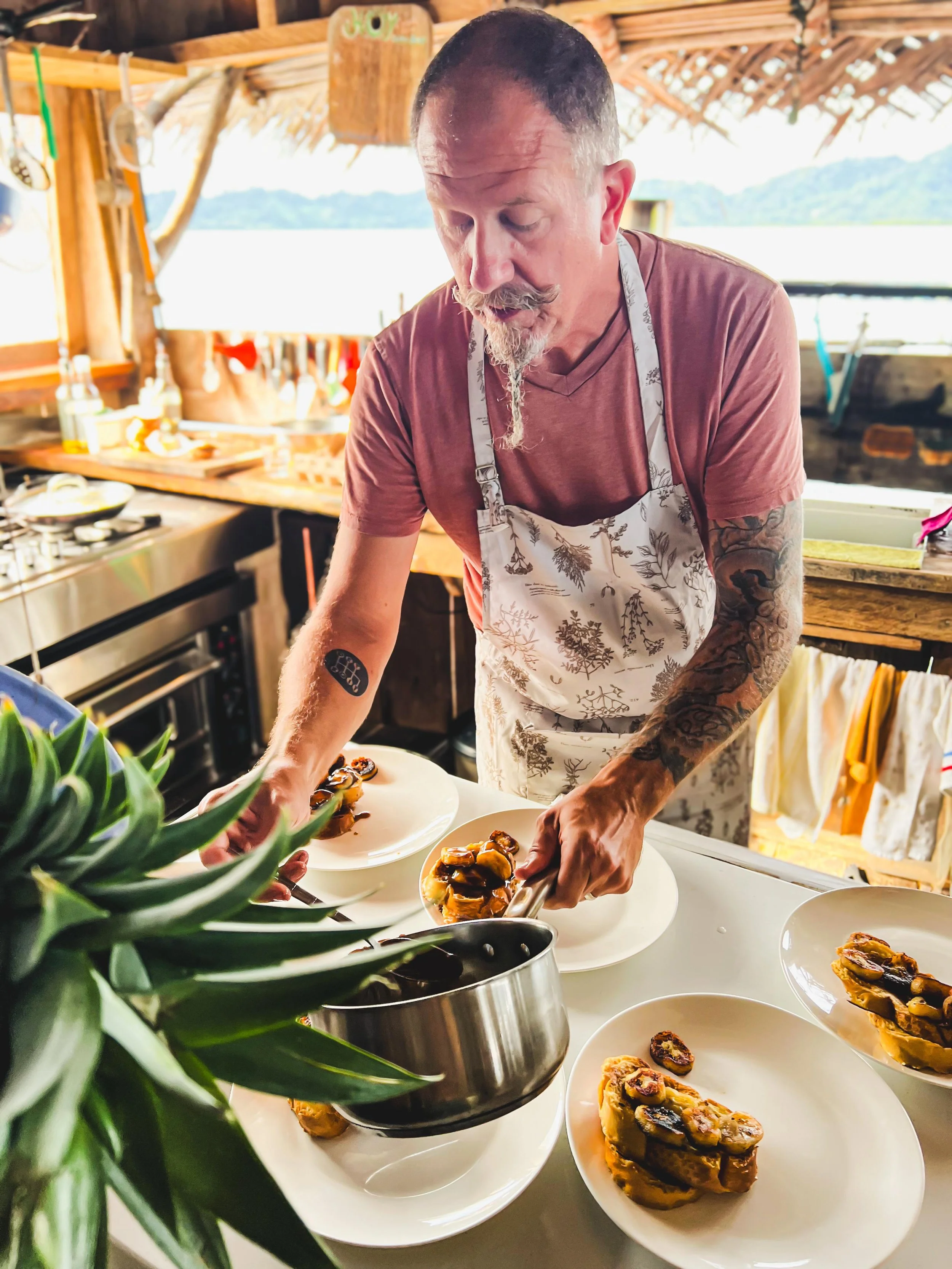 A man with short dark hair, tattoos on his arms, and a mustache and goatee, wearing a floral apron, is serving food on plates in a rustic kitchen with a view of water and mountains outside. There are plates of sliced eggplant on the table and a pineapple in the foreground.