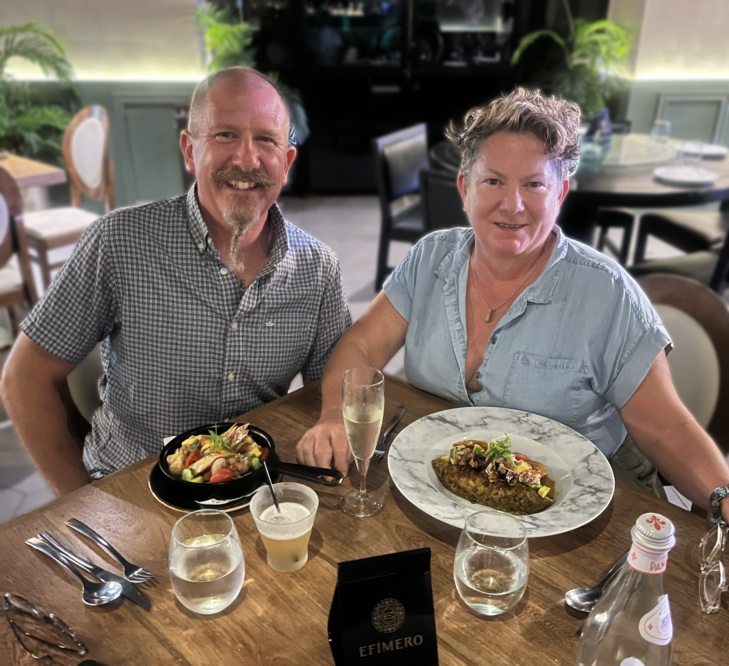 Two people sitting at a dining table with plates of food and glasses of white wine, smiling at the camera in a well-lit restaurant with plants in the background.