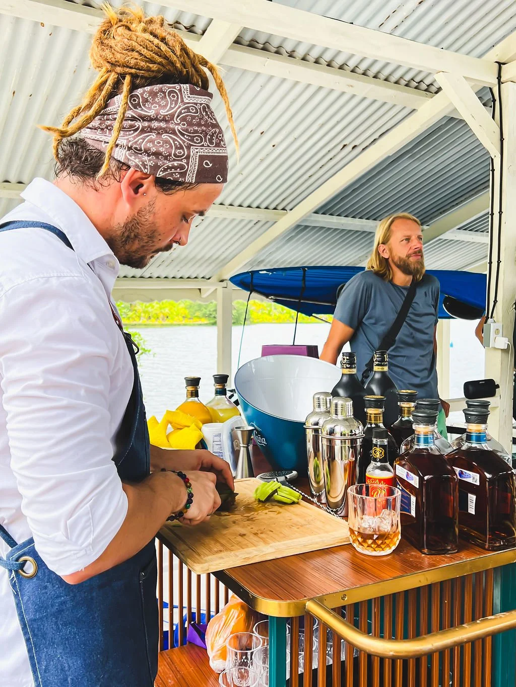 A bartender with dreadlocks wearing a bandana, white shirt, and apron is preparing a drink. Behind him is a man with long hair and a beard, leaning against the bar, which is outdoors by a body of water. The bar has bottles. The setting is under a cor