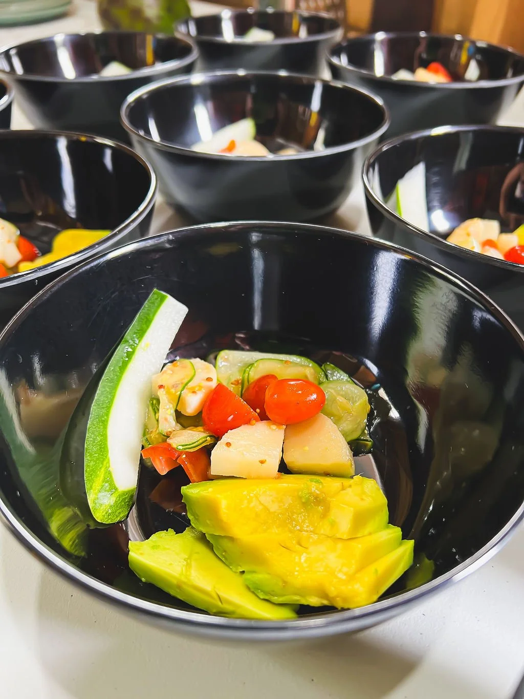 Black bowls filled with sliced avocado, cherry tomatoes, and cucumber, arranged on a white surface.