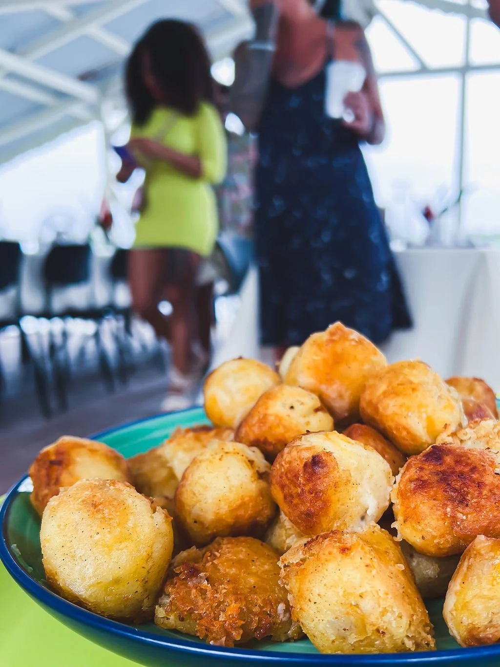 Close-up of a bowl of golden-brown fried cheese balls, with people in casual clothing and large windows in the background.