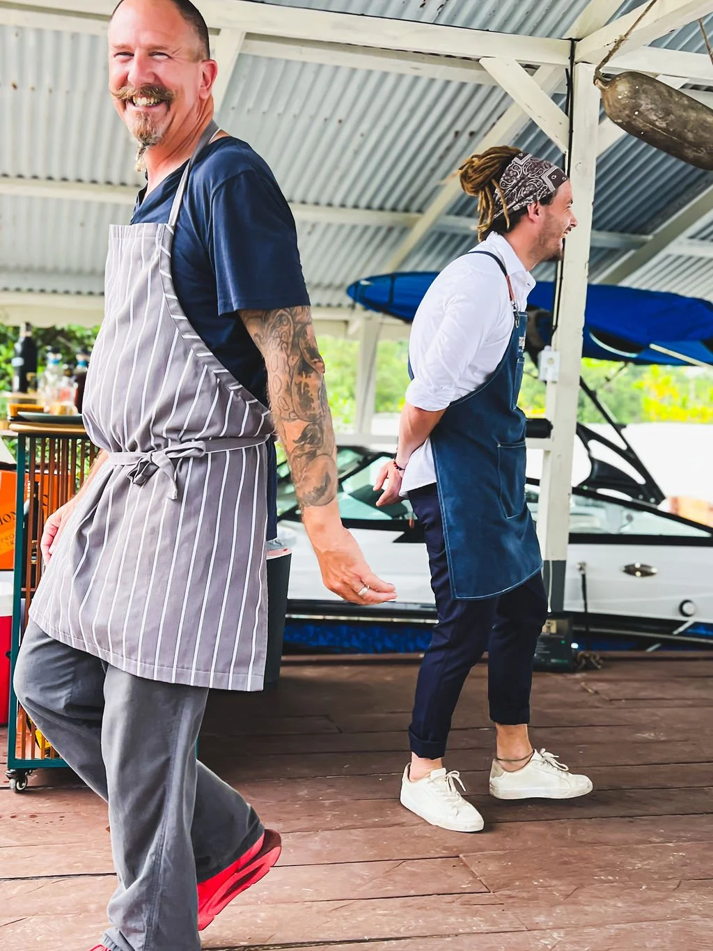 Two men wearing aprons and chef clothes, smiling and standing on a wooden deck under a metal roof at an outdoor food venue.