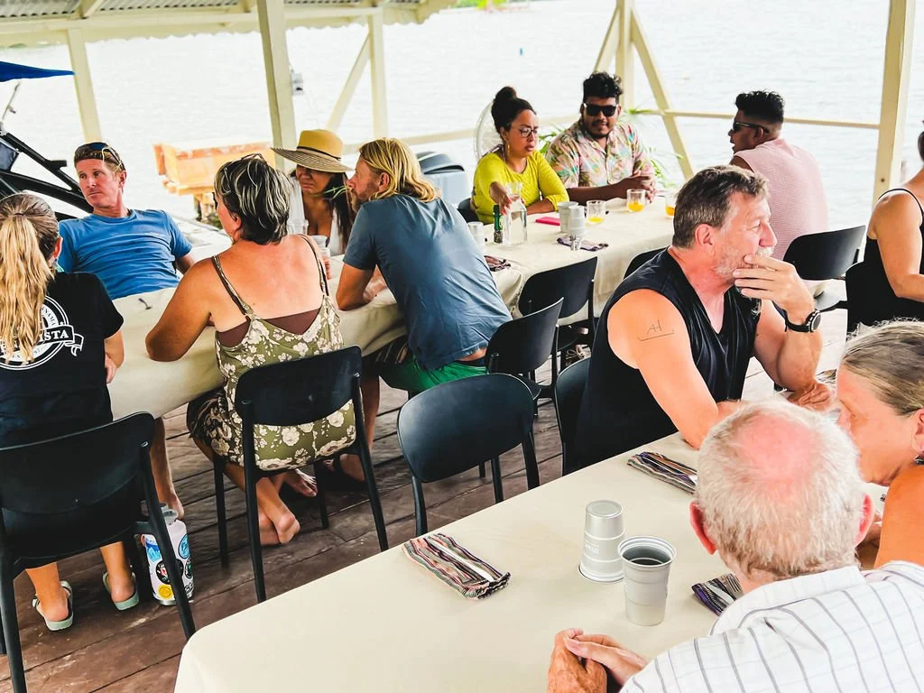 People sitting around a long table enjoying a meal at an outdoor waterfront setting.