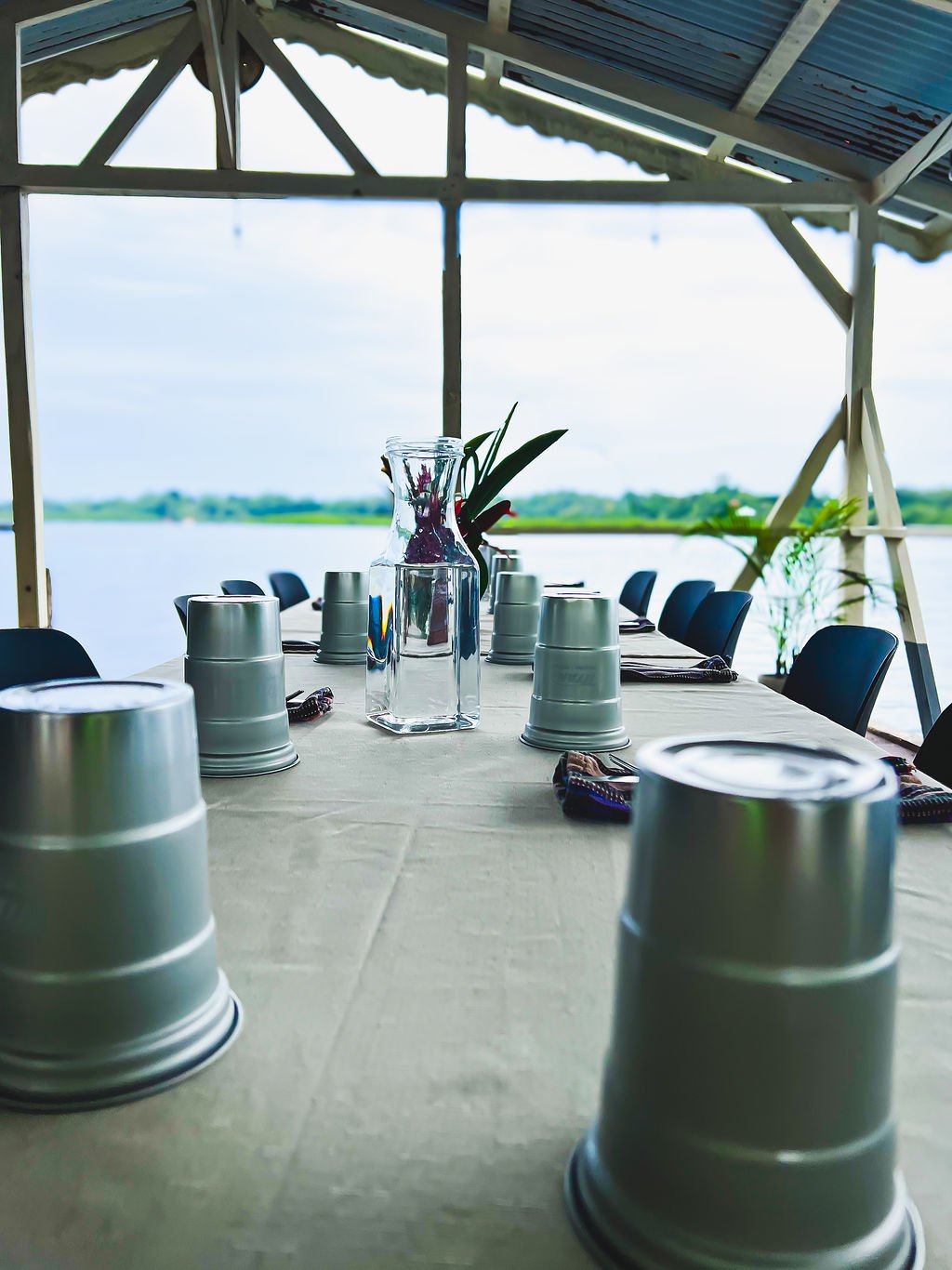 Long dining table set with metal cups, a glass pitcher with flowers, and a vase with plants, overlooking a body of water under a roofed outdoor pavilion.