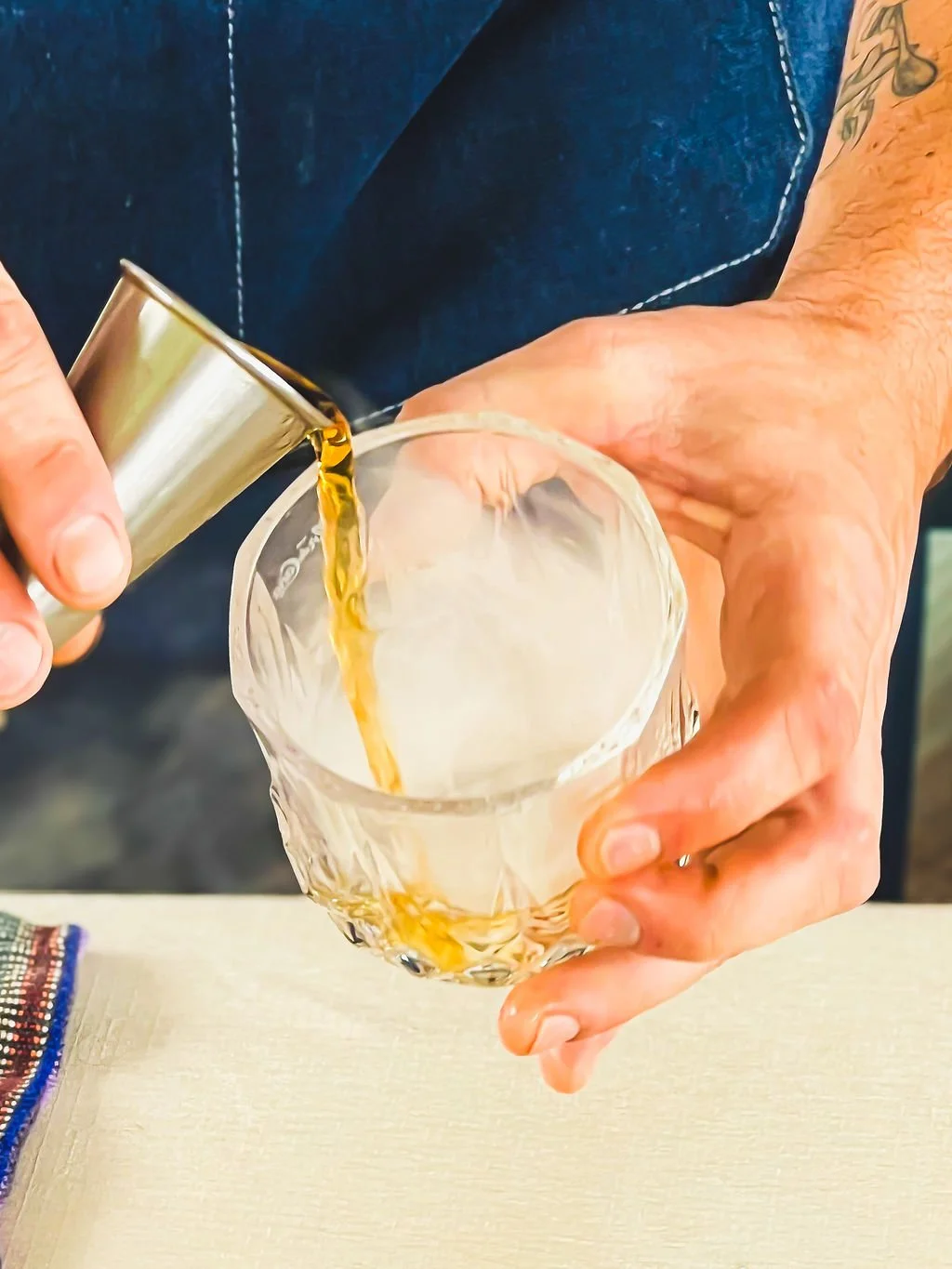 Person pouring a beverage from a metal shaker into a glass. Person's hands and part of arm visible, wearing a dark sleeveless shirt.