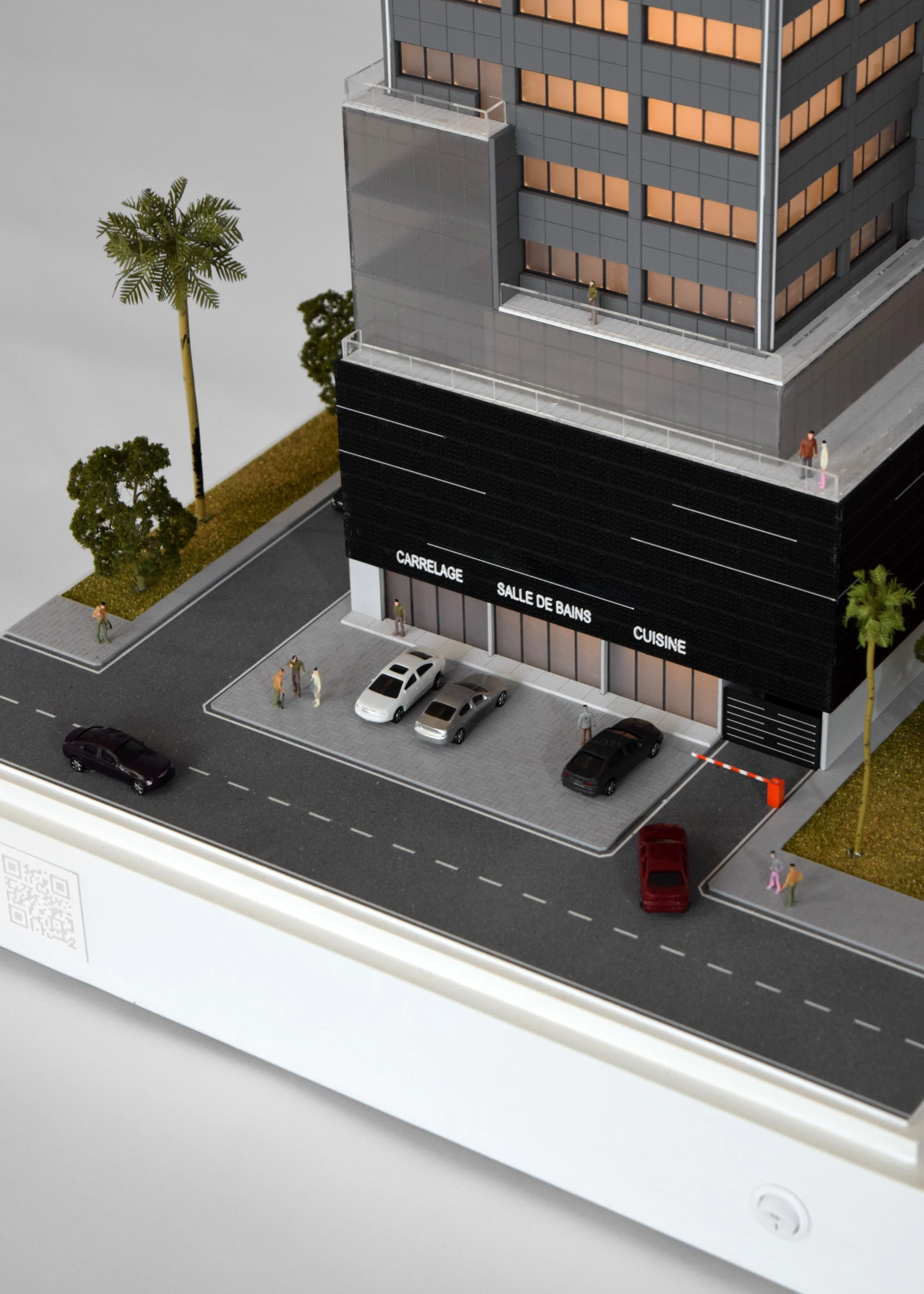 Model of a modern multi-story building with a parking lot, trees, and small figures of people walking outside. The building has a black signage area with white text in French indicating a tile store, a bathroom, and a kitchen.
