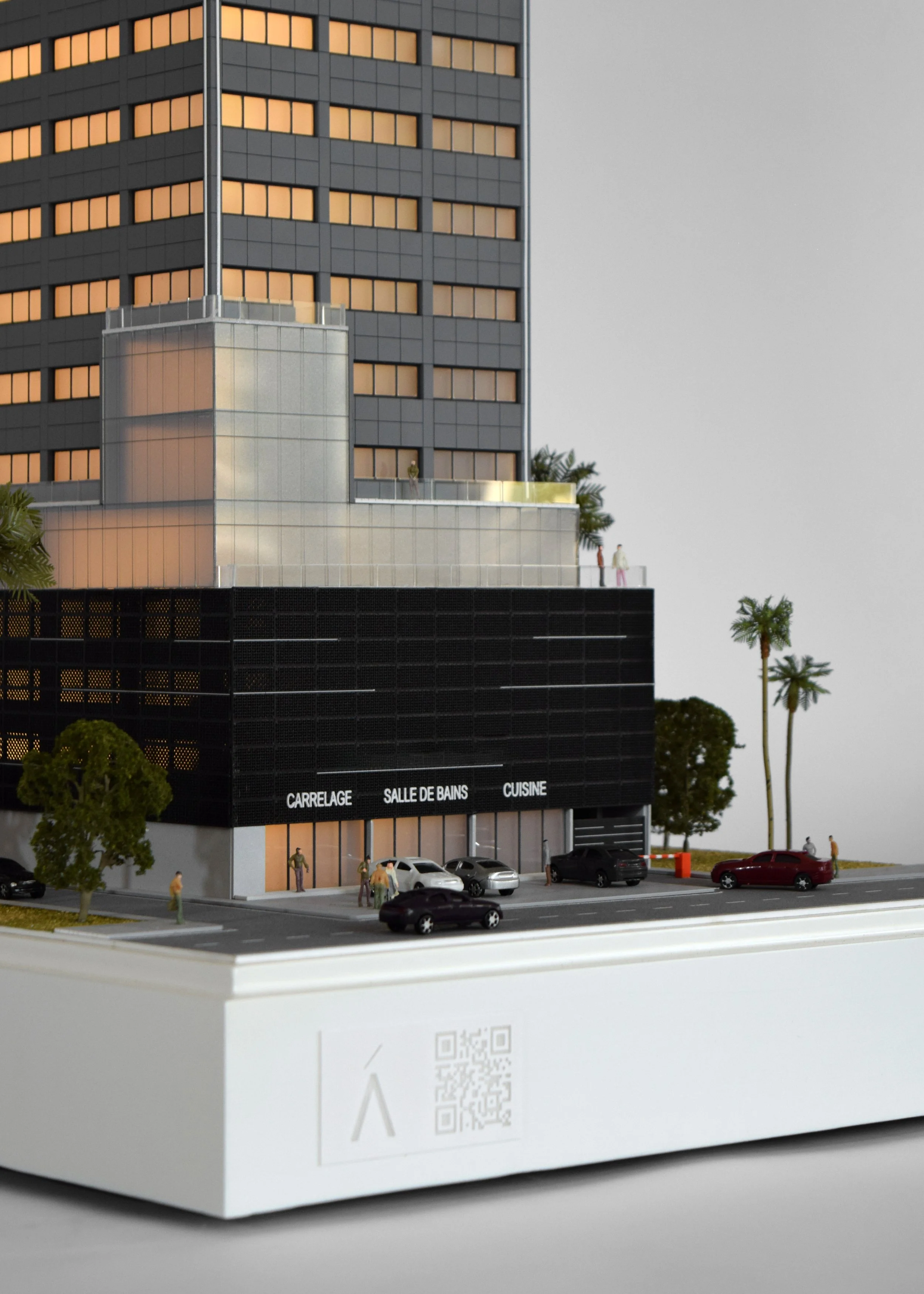 A detailed architectural model of a modern multi-story building with a ground-floor commercial space labeled "CARRELAGE," "SALLE DE BAINS," and "CUISINE." The building features a black facade and large windows, with small scaled figures and cars in the surrounding area, indicating an urban setting.