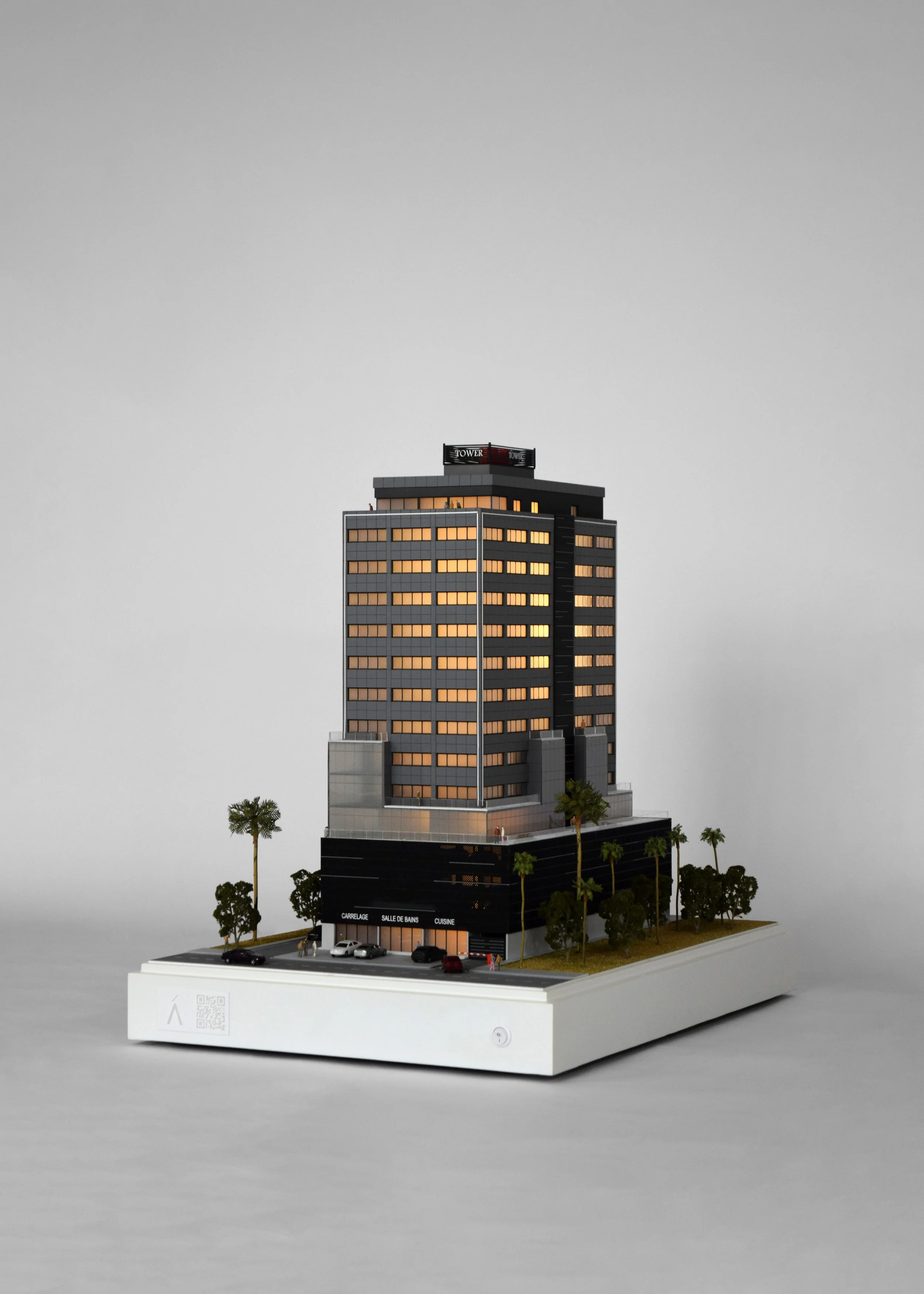 Model of a modern high-rise building with a parking lot, trees, and a gray sky background.