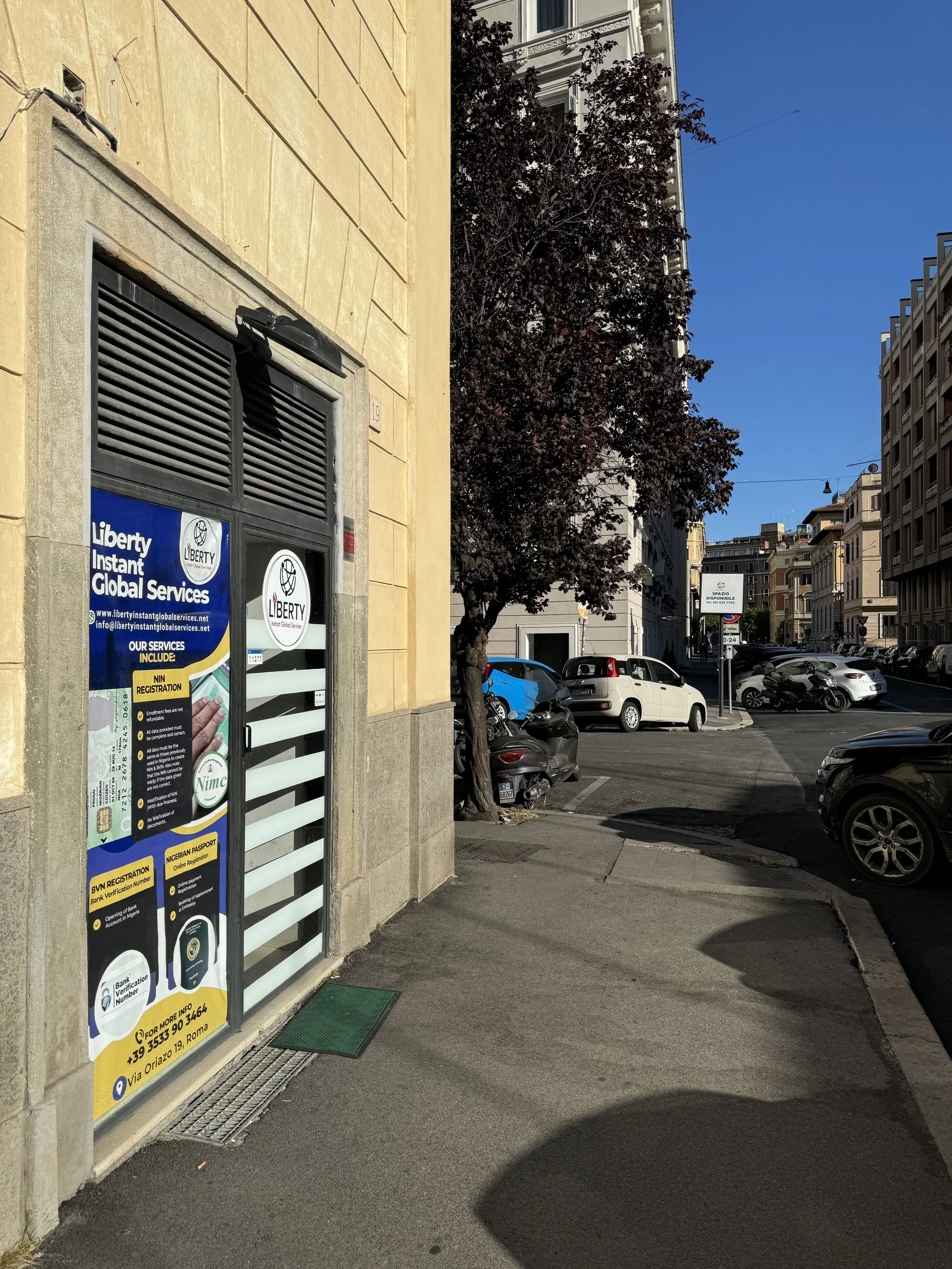 A city street with parked cars and motorcycles, a yellow building on the left with a sign for Liberty Instant Global Services, and a tree with dark leaves. The sky is clear and blue.