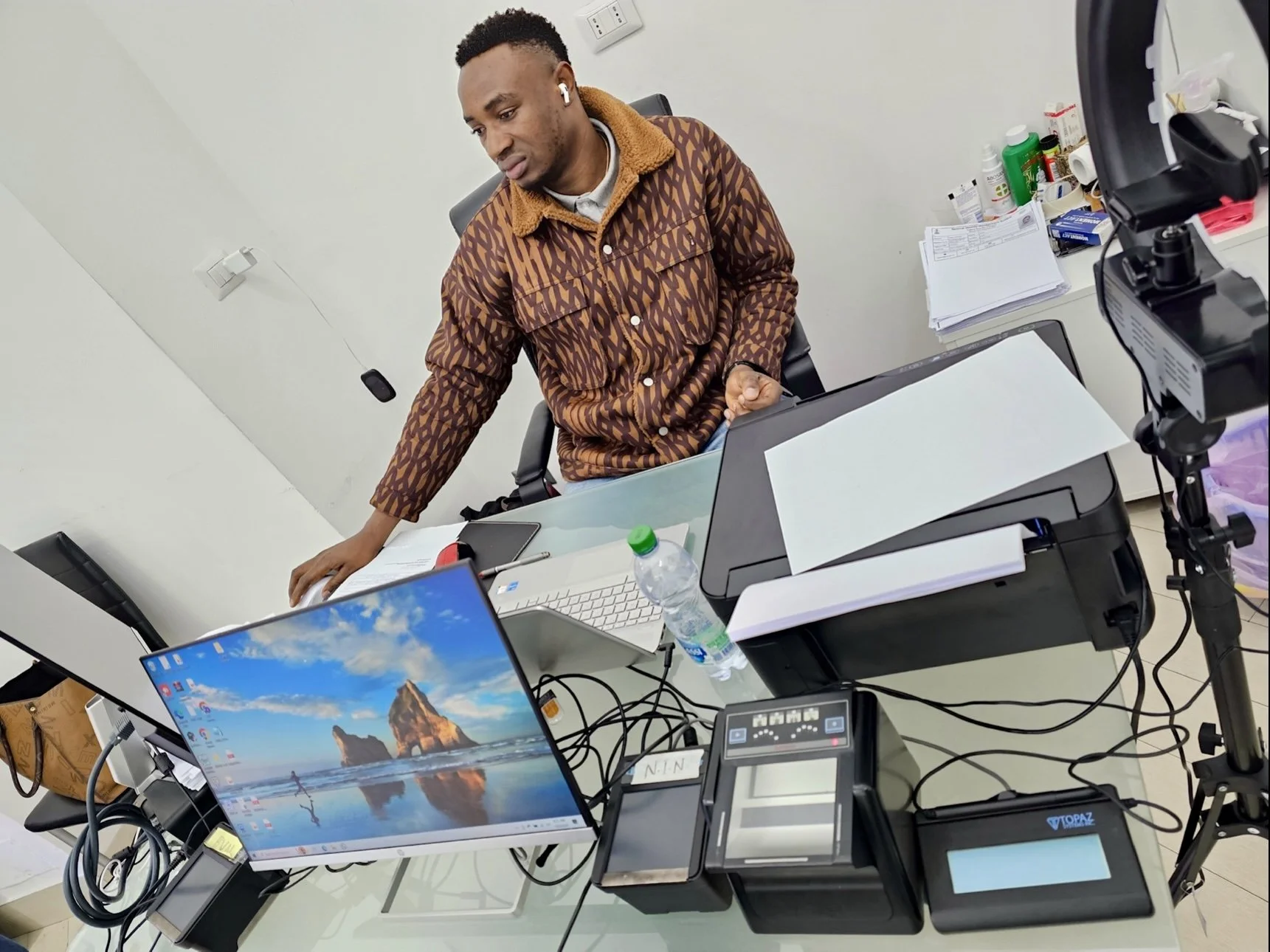 Man in a patterned brown jacket standing at a cluttered desk with multiple electronic devices, papers, and supplies in an office.