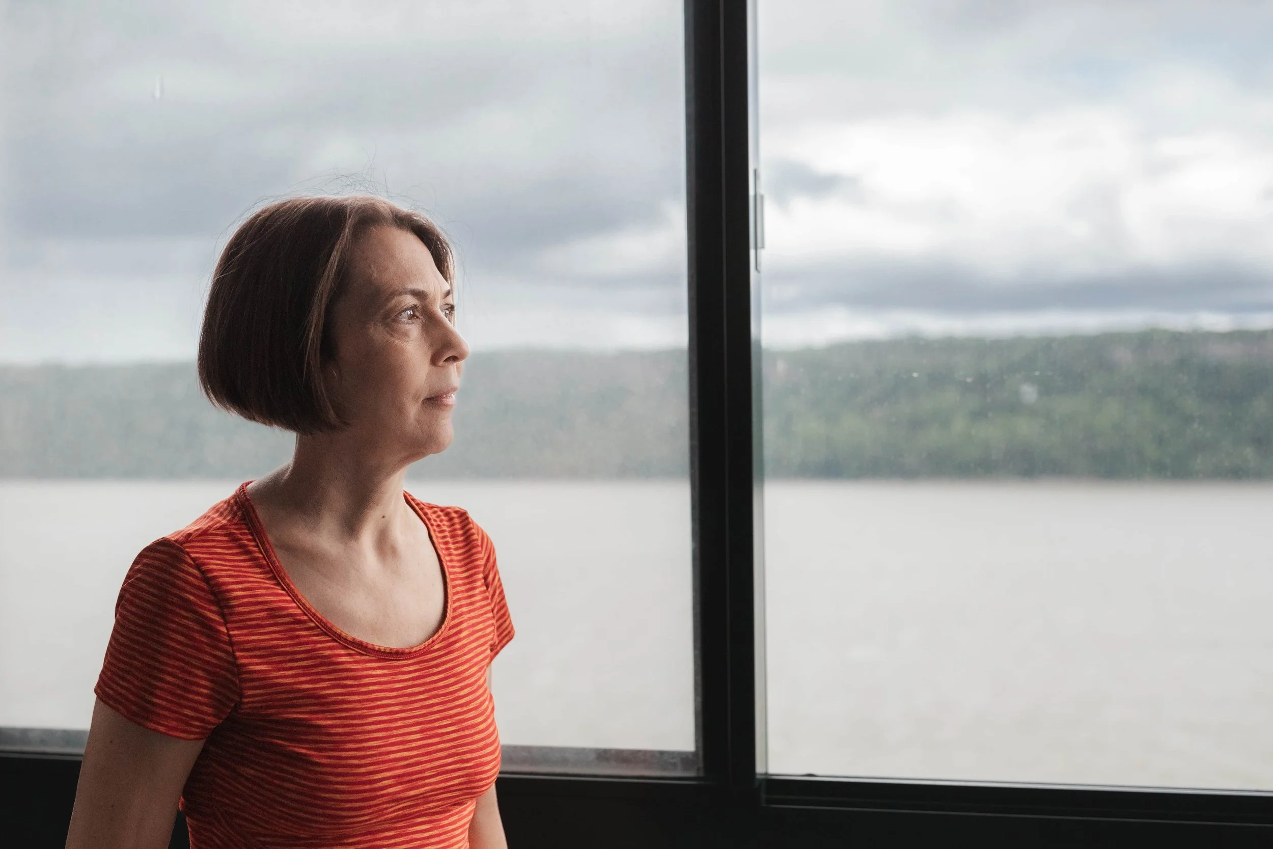 A woman with short brown hair looking out a large window with a cloudy sky and landscape outside.