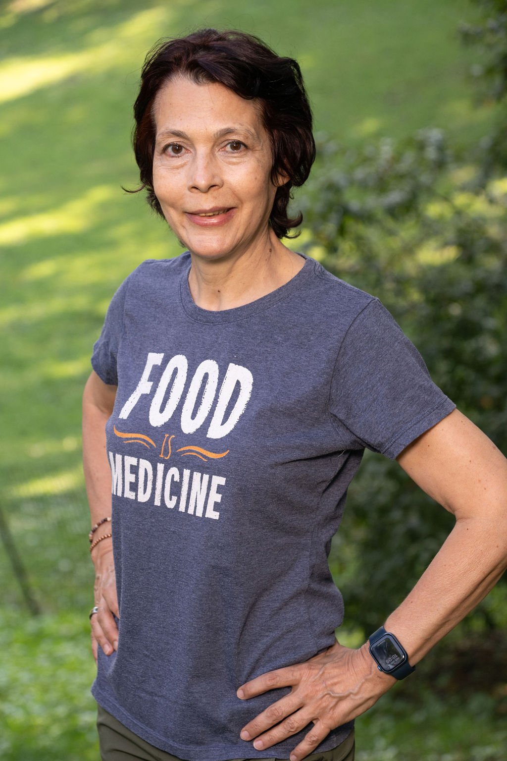 A woman outdoors with short dark hair, wearing a dark gray T-shirt with the slogan 'Food is Medicine,' and an Apple Watch, standing with her hand on her hip against a blurred green background.