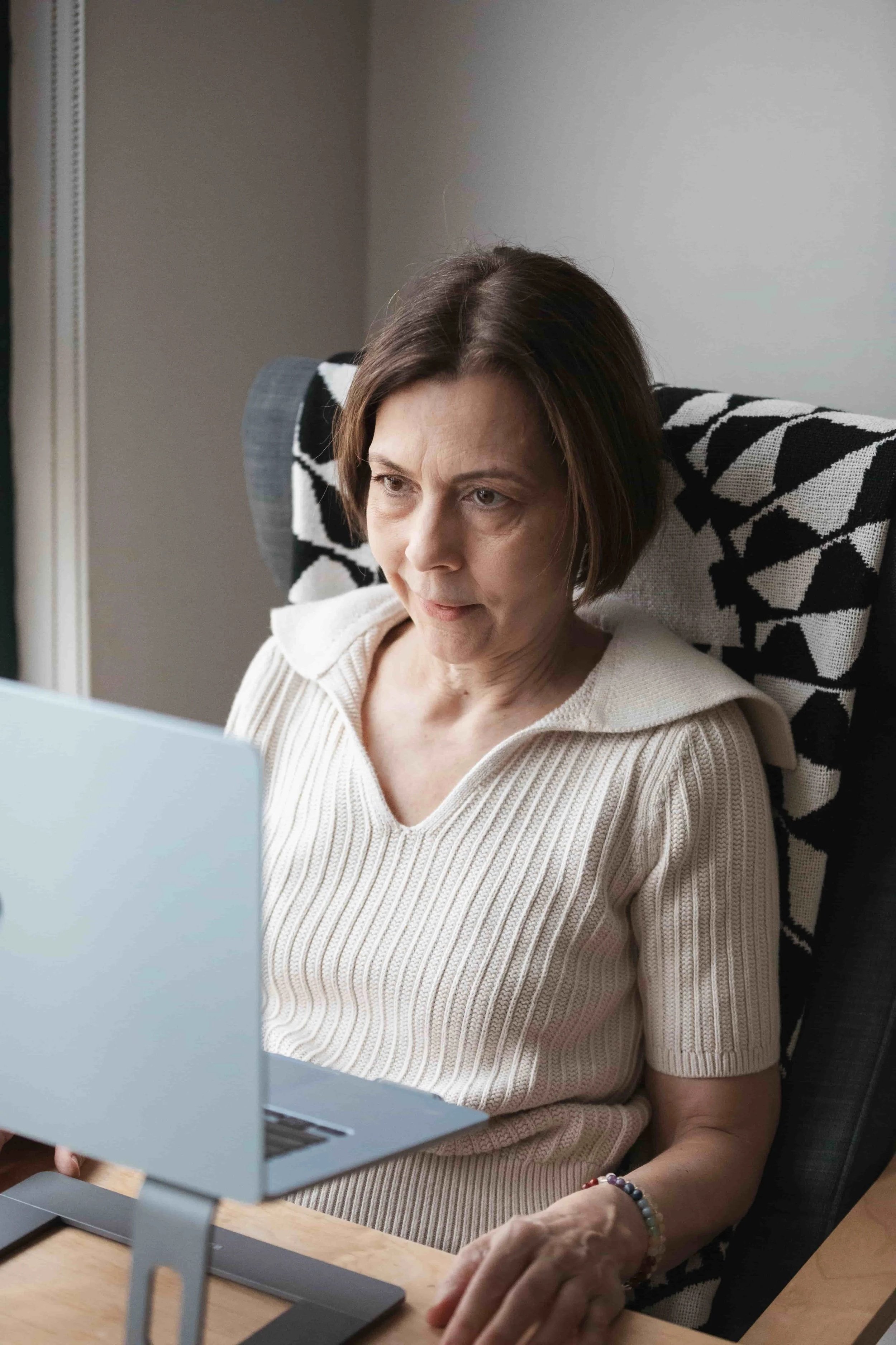 A woman with short brown hair wearing a beige knitted sweater working on a laptop at a wooden desk. She is seated in a black and white patterned chair with a gray backrest, in a room with a gray wall.