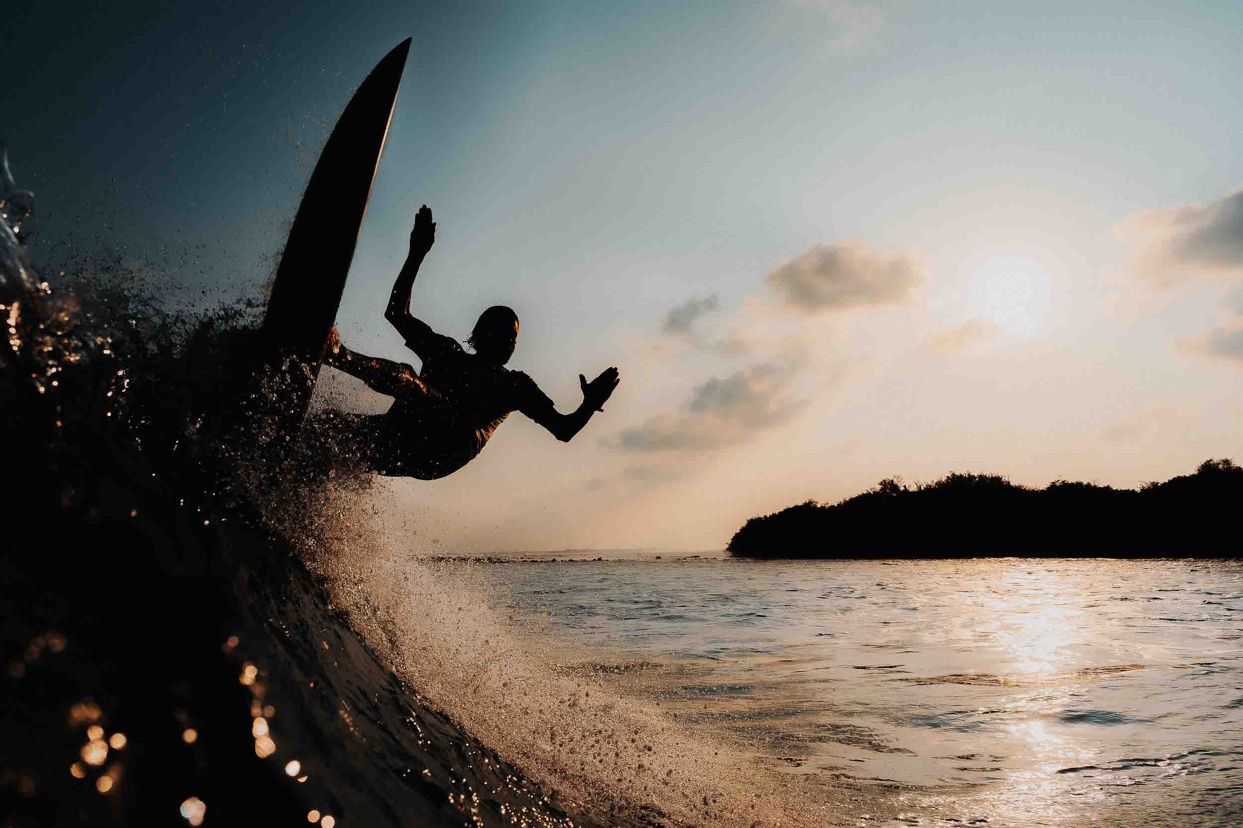 Silhouette of a person falling off a surfboard into the water during sunset, with waves and a distant shoreline.