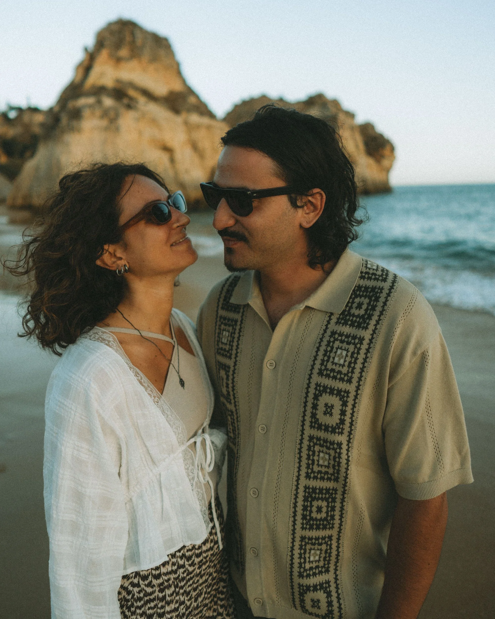 Romantic close-up portrait of a couple on the beach at sunset in Algarve, warm light illuminating their faces with ocean and cliffs behind.