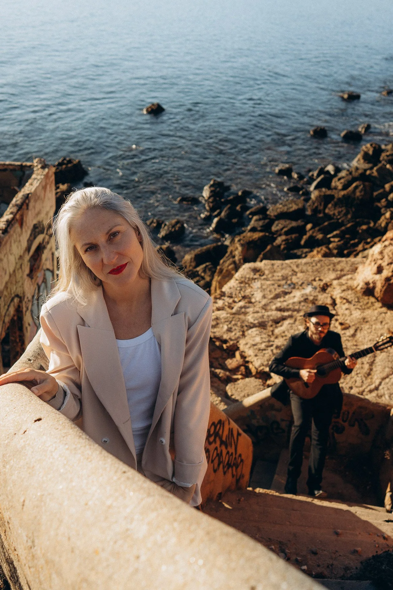 A woman with blonde hair and red lipstick leaning on a concrete barrier near the water, with a man playing guitar in the background on rocky steps.