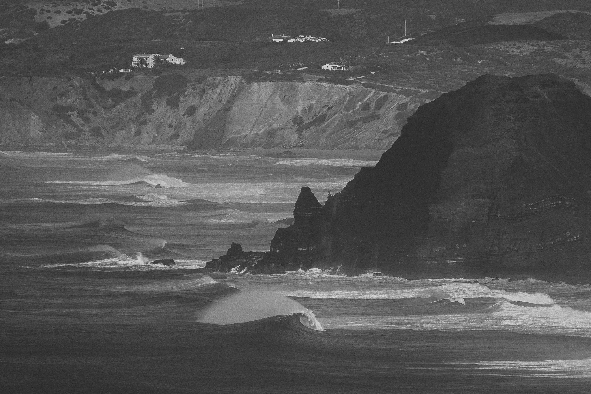 Black and white photo of a rugged coastline with large cliffs and waves crashing against the rocks, with houses on top of the cliffs in the background.