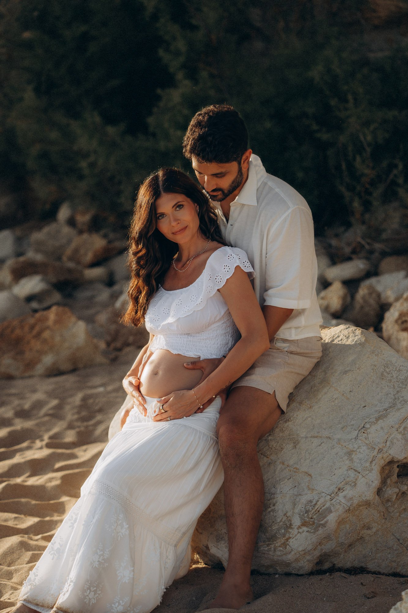Couple holding baby bump sitting on rock at golden hour, intimate maternity photography Algarve