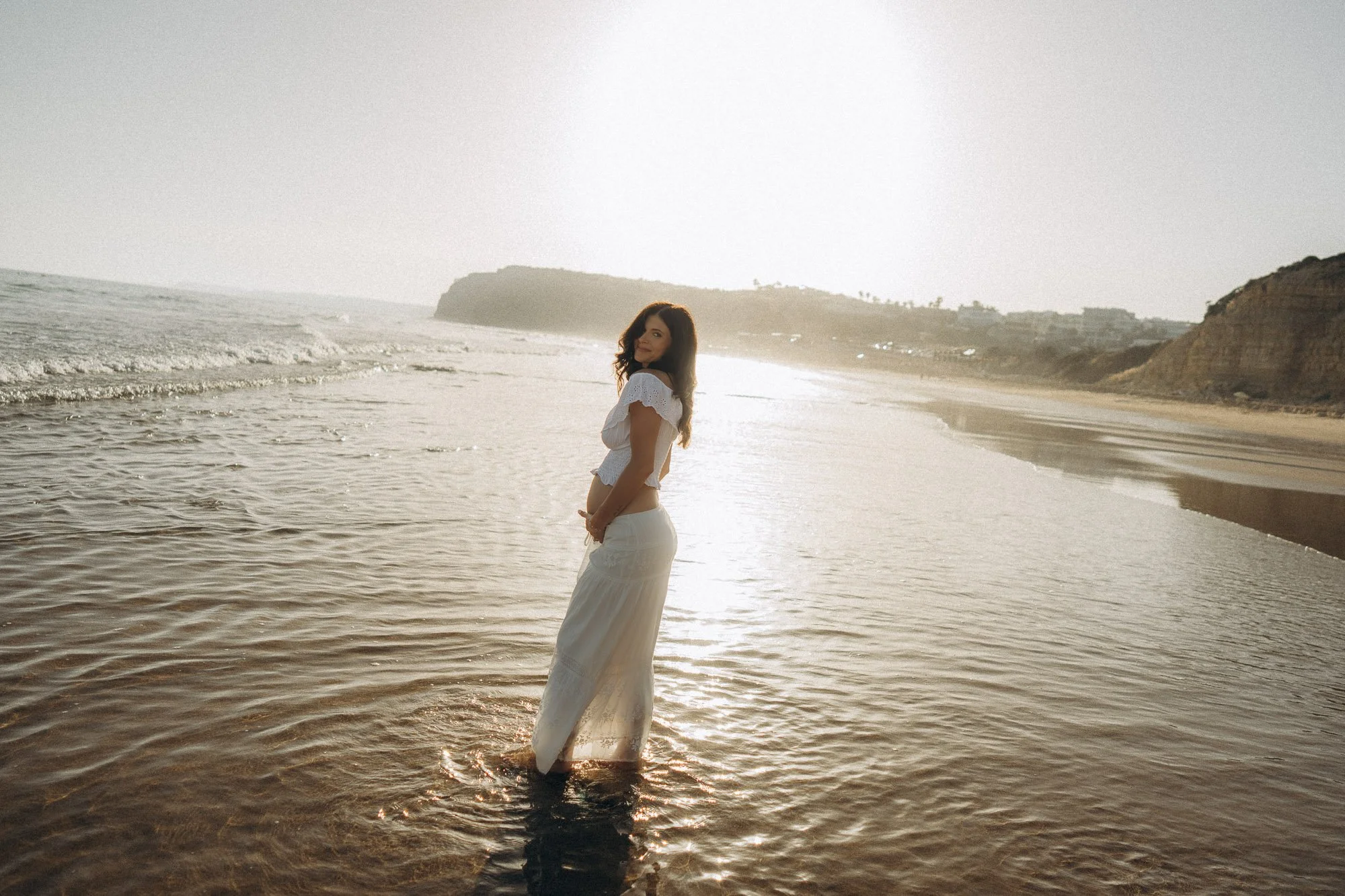 Maternity photographer Algarve, pregnant woman standing in shoreline water at golden hour, coastal cliffs