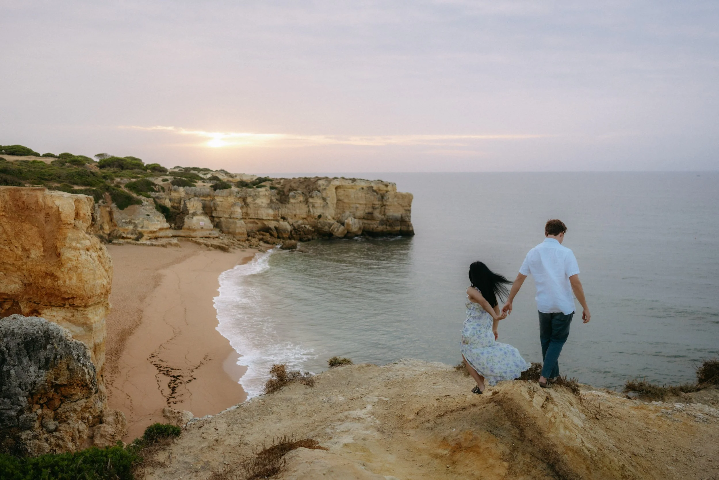 Walking together toward sunset light, soft tones hitting the limestone formations along the Algarve coast.