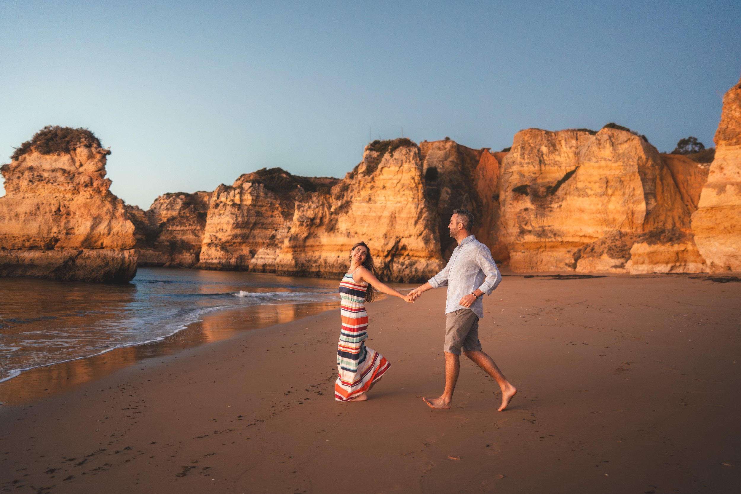 Romantic couple running barefoot on Praia Dona Ana at sunset, golden light reflecting on the cliffs of the Algarve.