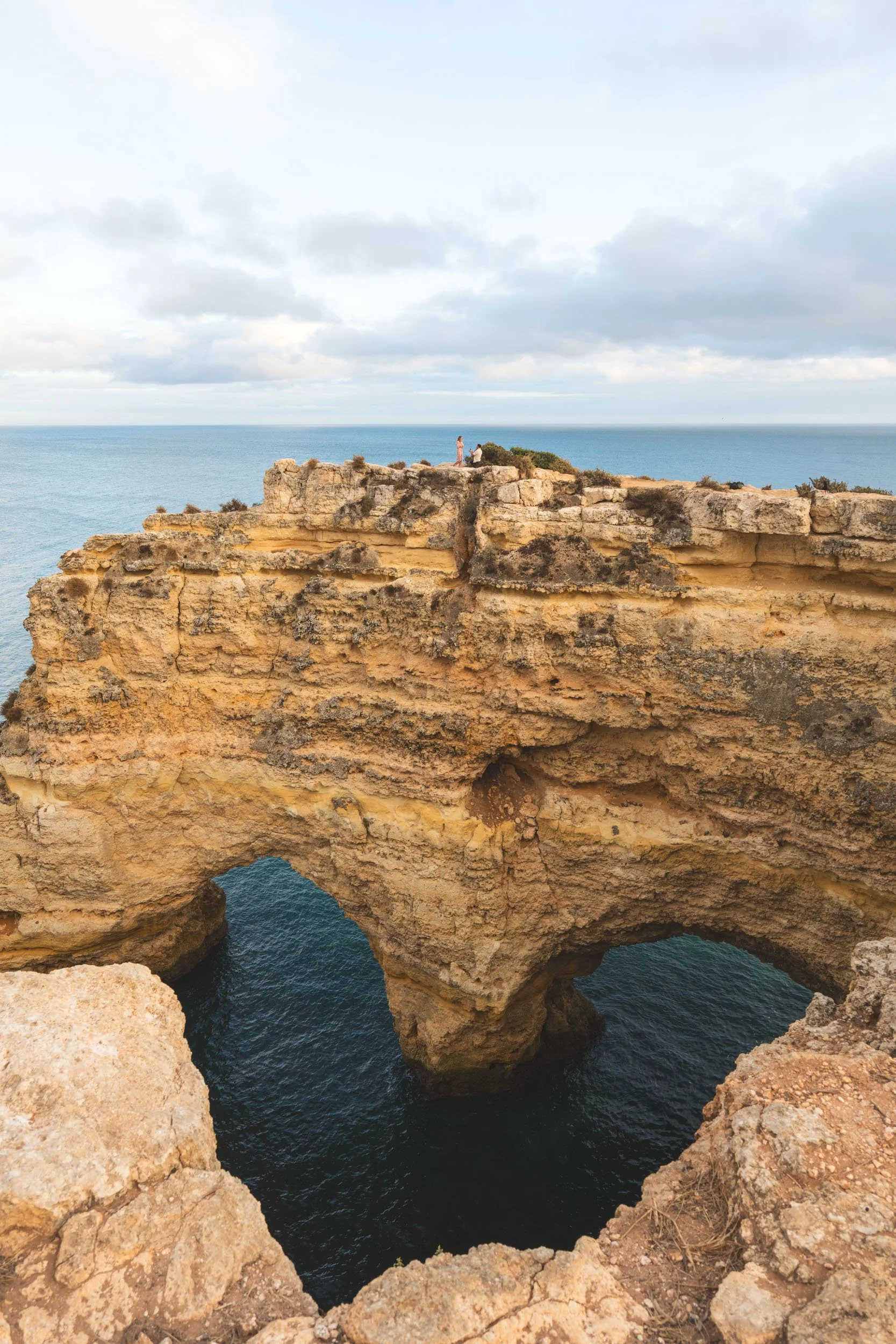 Wide view of a couple on the iconic Praia da Marinha cliffs in the Algarve, photographed above the natural rock arch during a romantic engagement session.