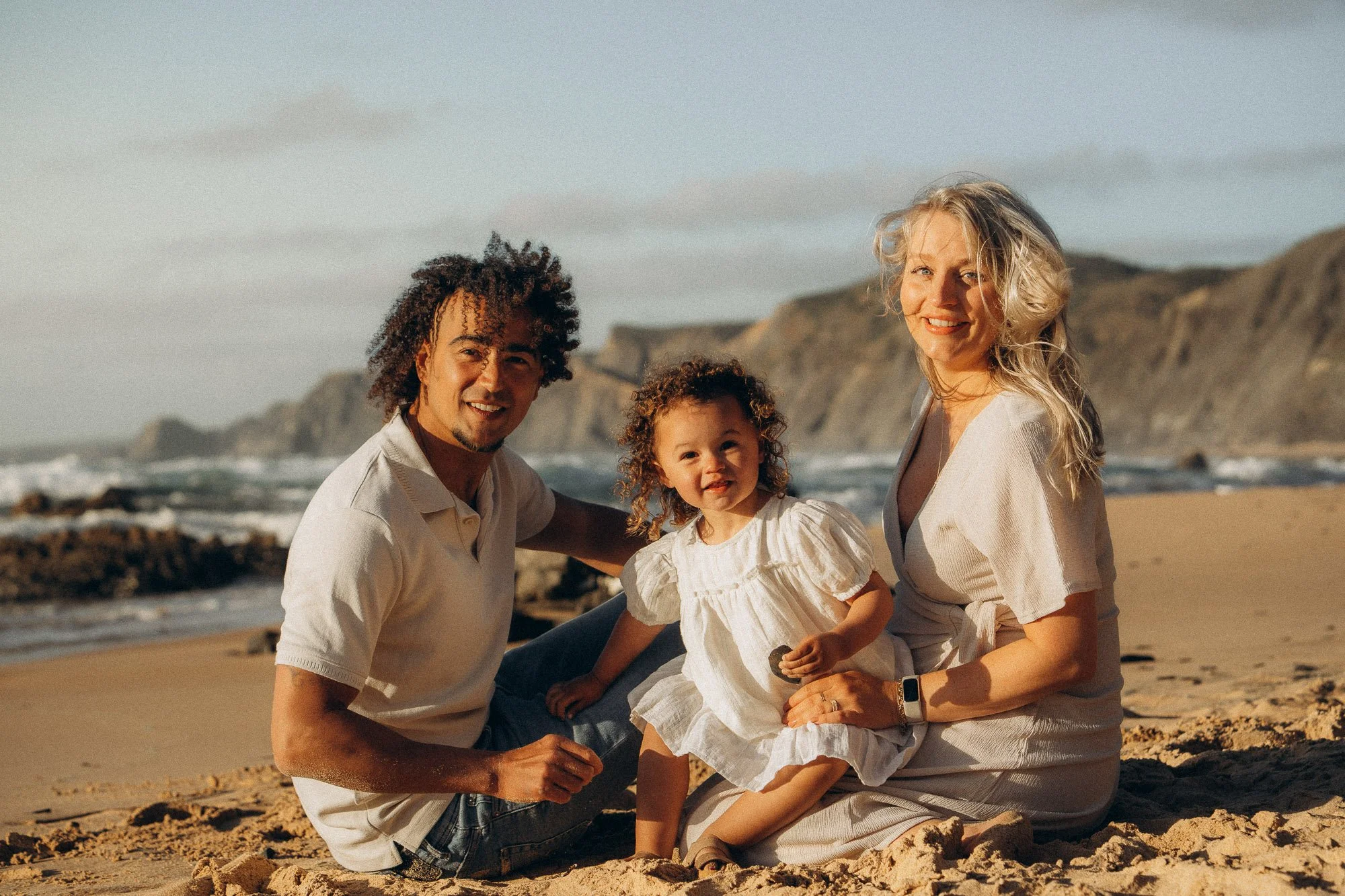 Family photography session on Algarve beach at golden hour, couple sitting on sand with toddler daughter, cliffs in background