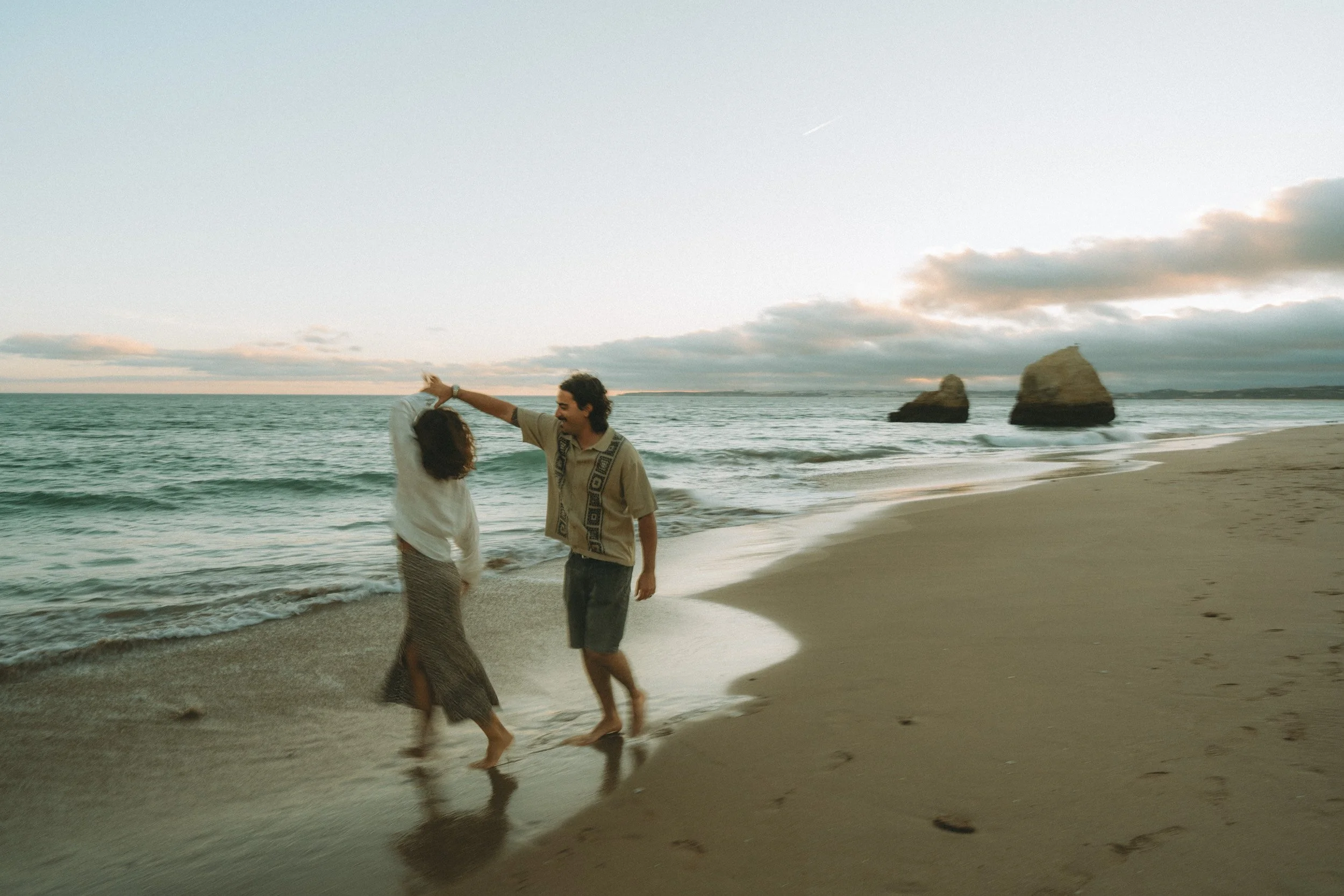 Wide scenic portrait of a couple seated on the sand at Praia dos Três Irmãos, framed by ocean and iconic rock stacks.