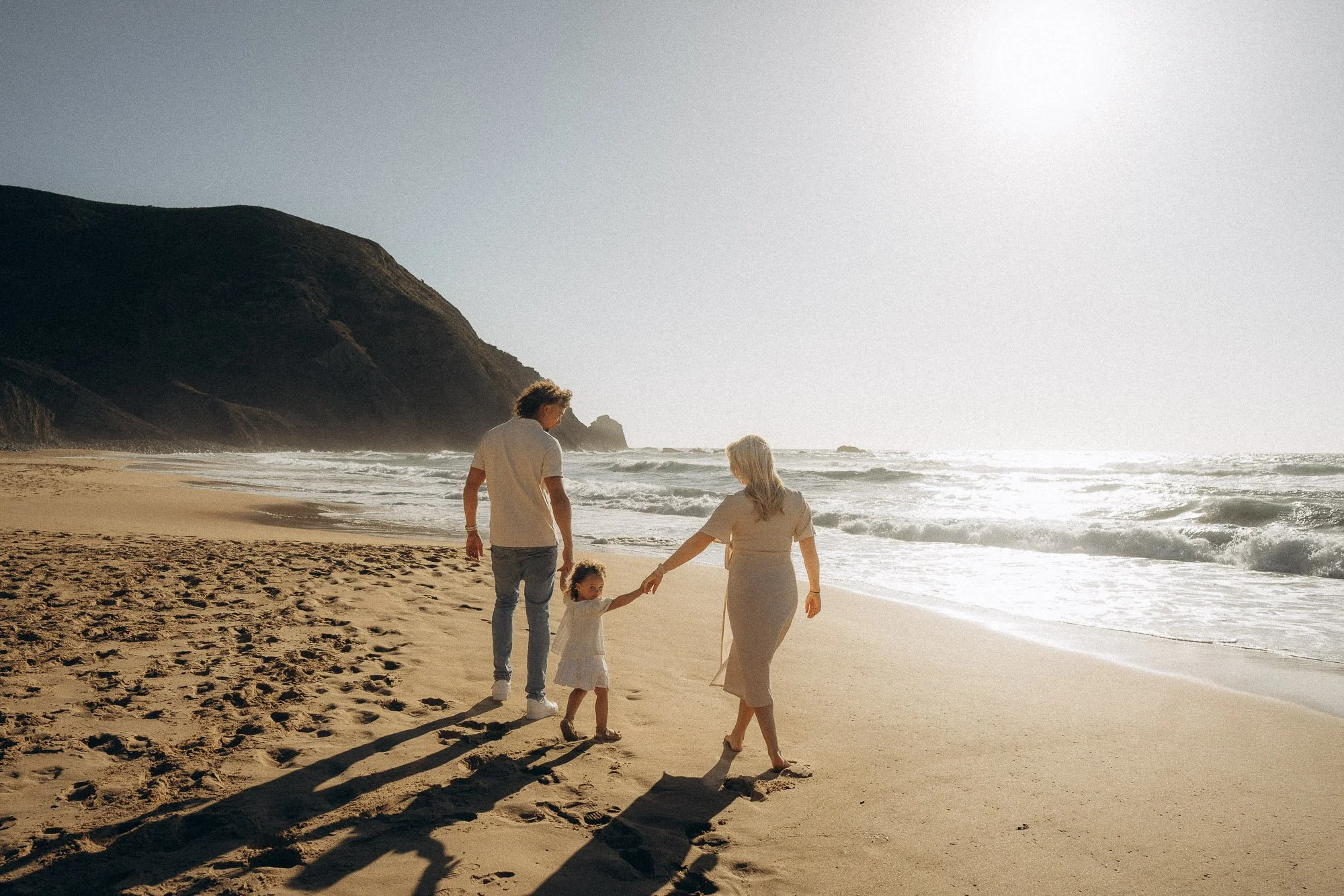 Family of three walking hand in hand along Algarve shoreline, natural light family photography Portugal