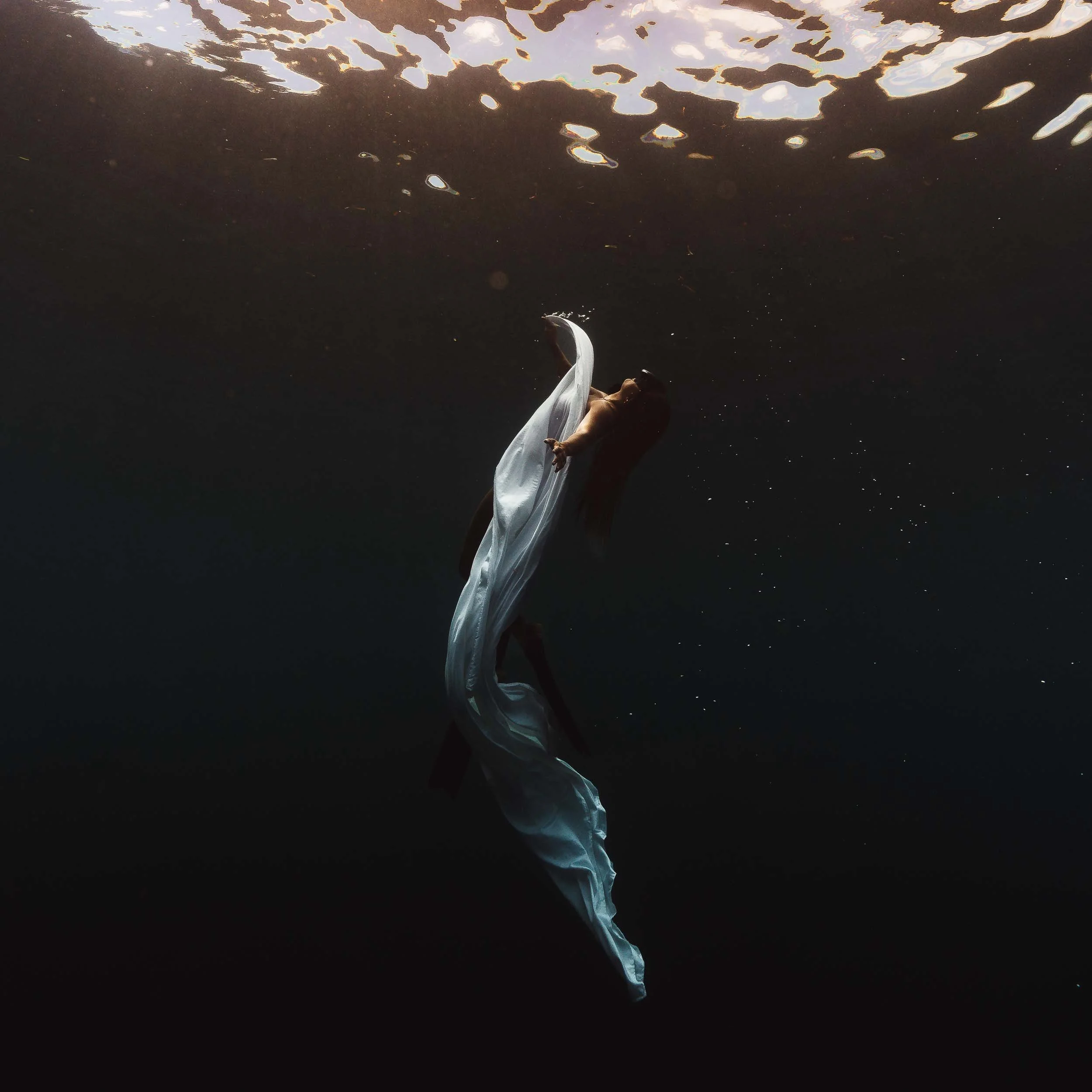 A person underwater, wearing a flowing white dress, with their head tilted back and long hair floating. The photo is taken from beneath the surface, with the water surface visible at the top.