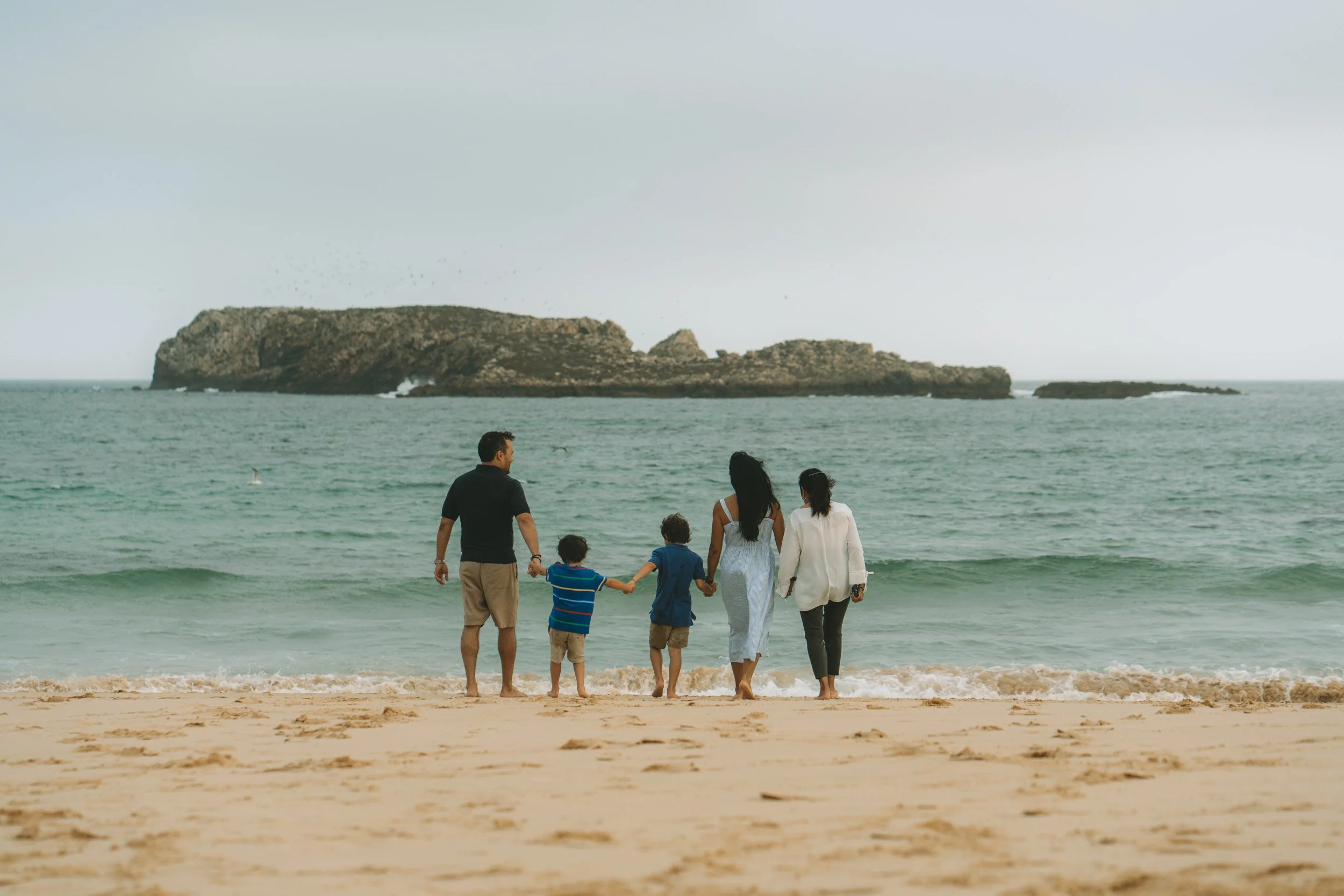 Family of six holding hands and walking into the ocean at the beach with a rocky island in the background. Praia do Martinhal, Sagres