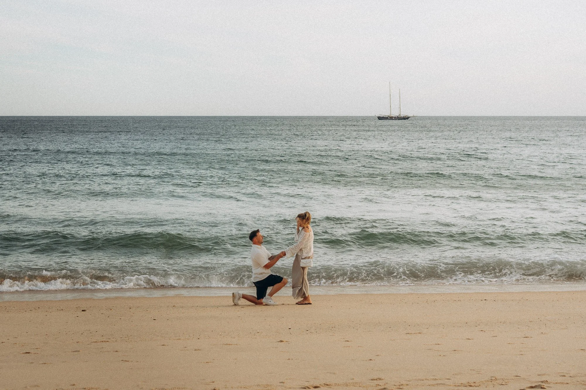 Surprise beach proposal Algarve, man on one knee at shoreline with sailboat in background, proposal photographer Portugal