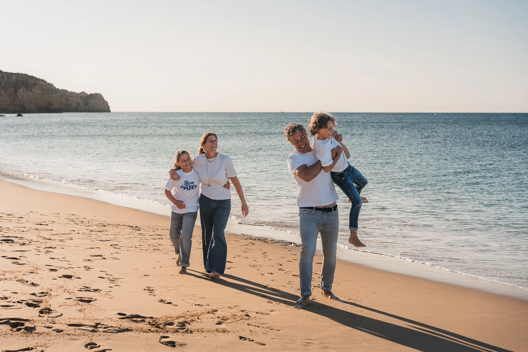 Family of 4 laughing and holding hands while strolling along the Porto de Mós shore in Lagos, Algarve.