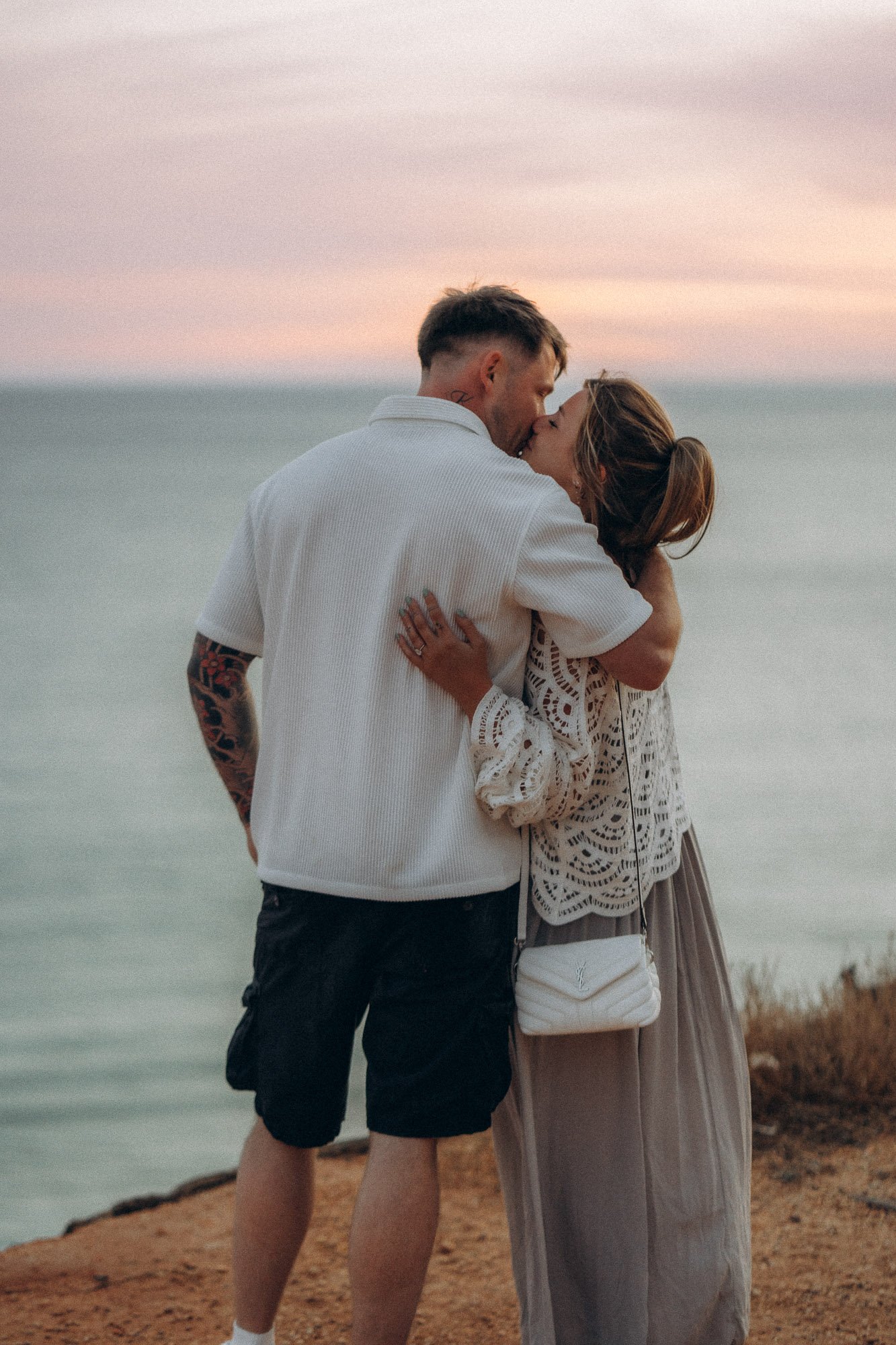 Couple kissing on Algarve cliff at dusk with pastel sky and ocean, engagement photography Portugal