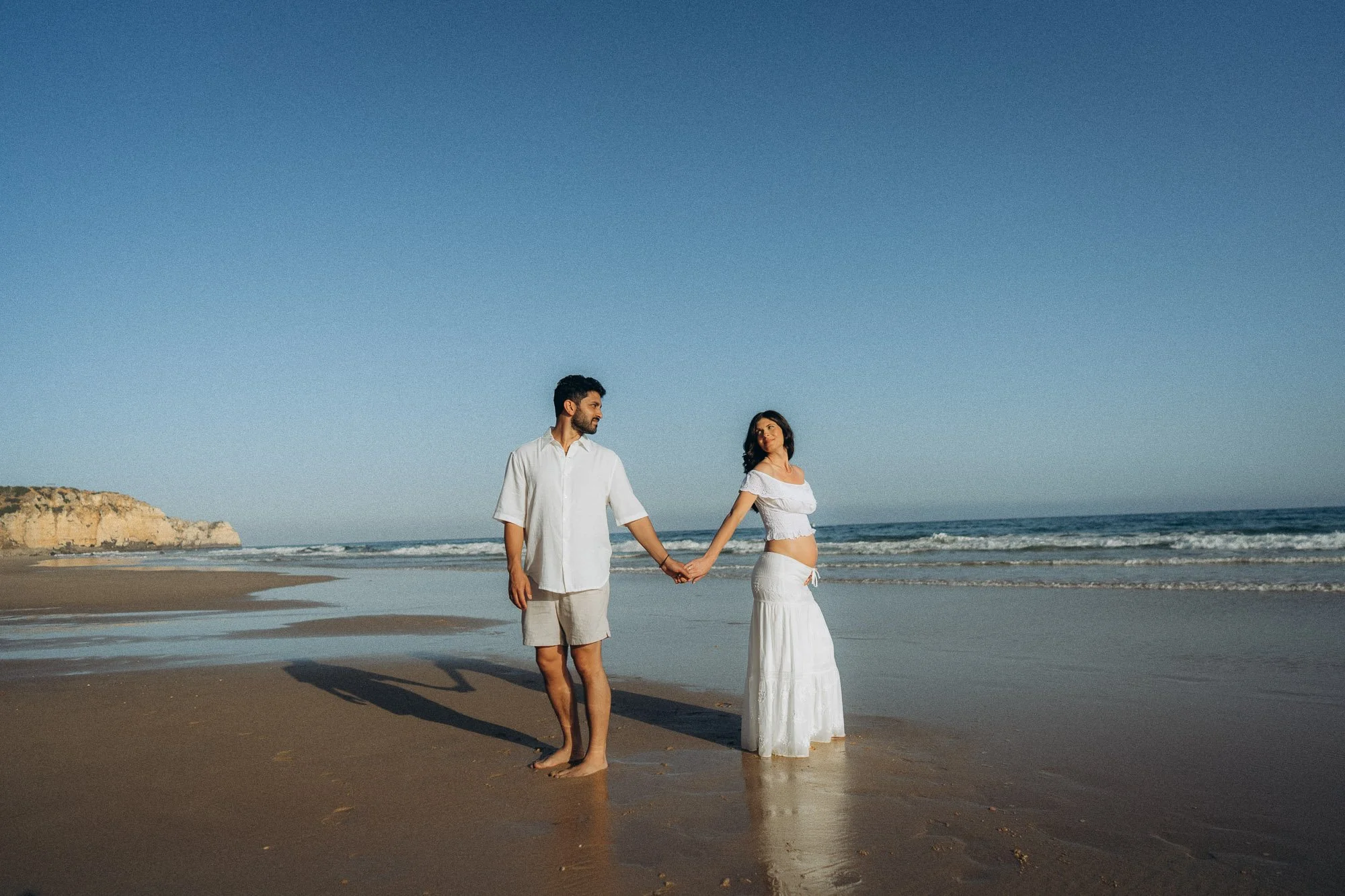 Maternity session on Algarve beach, pregnant couple holding hands at shoreline with cliffs in distance