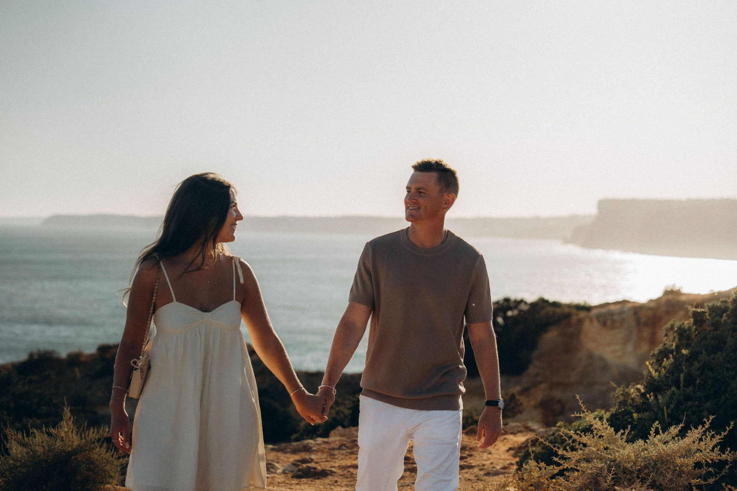 Candid walking shot with dramatic coastline behind. Natural interaction, no forced posing.