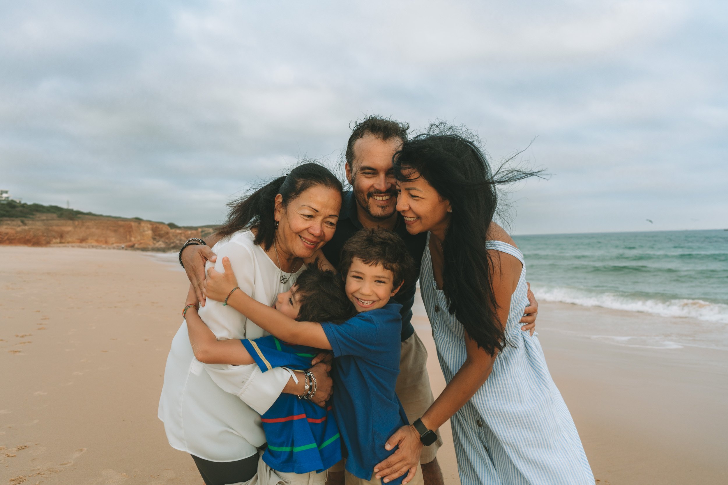 A family of four hugging and smiling on a beach, with the ocean and cloudy sky in the background. Algarve family photographer